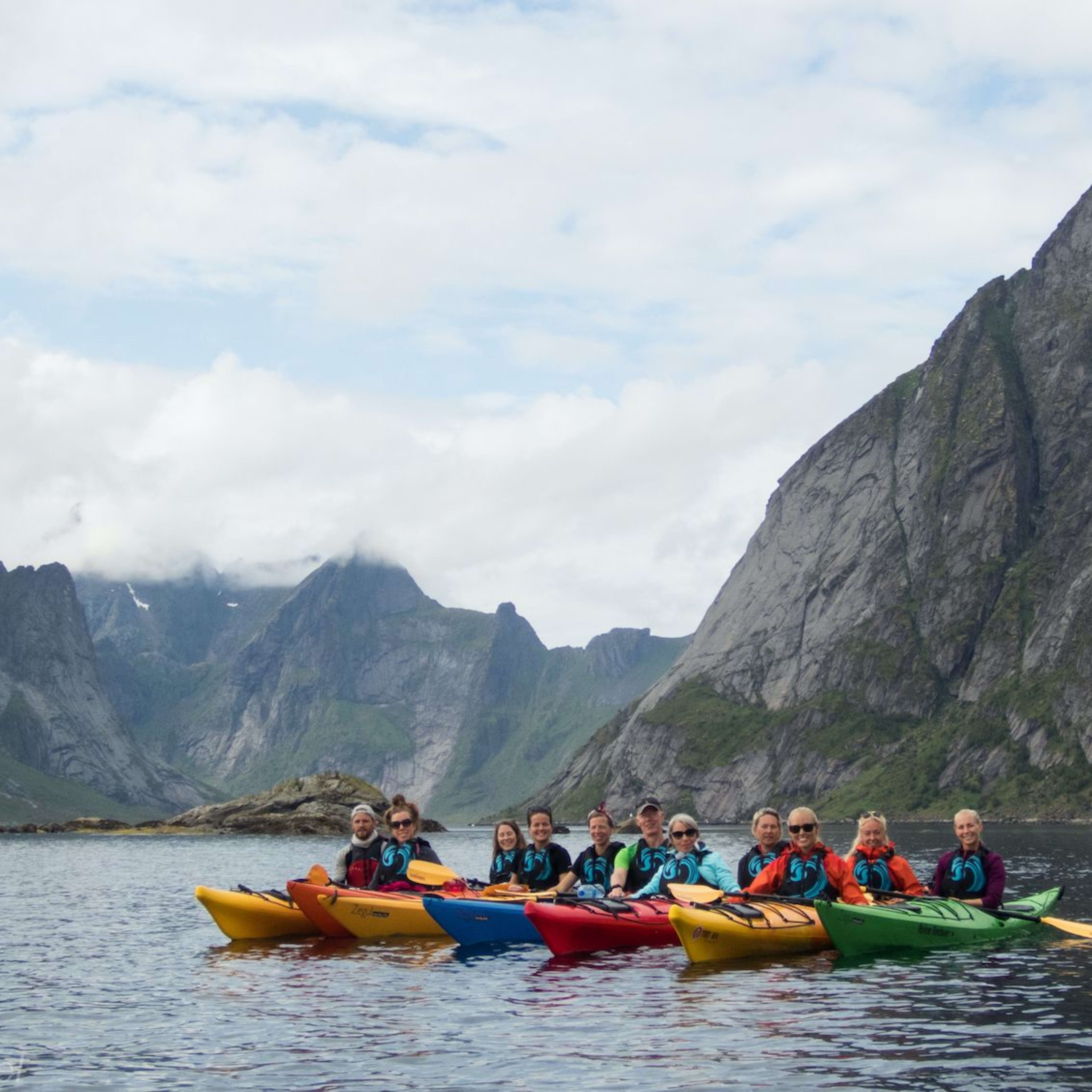 Group of kayakers in colorful kayaks on calm water, with jagged mountain peaks rising and cloudy skies in the background.