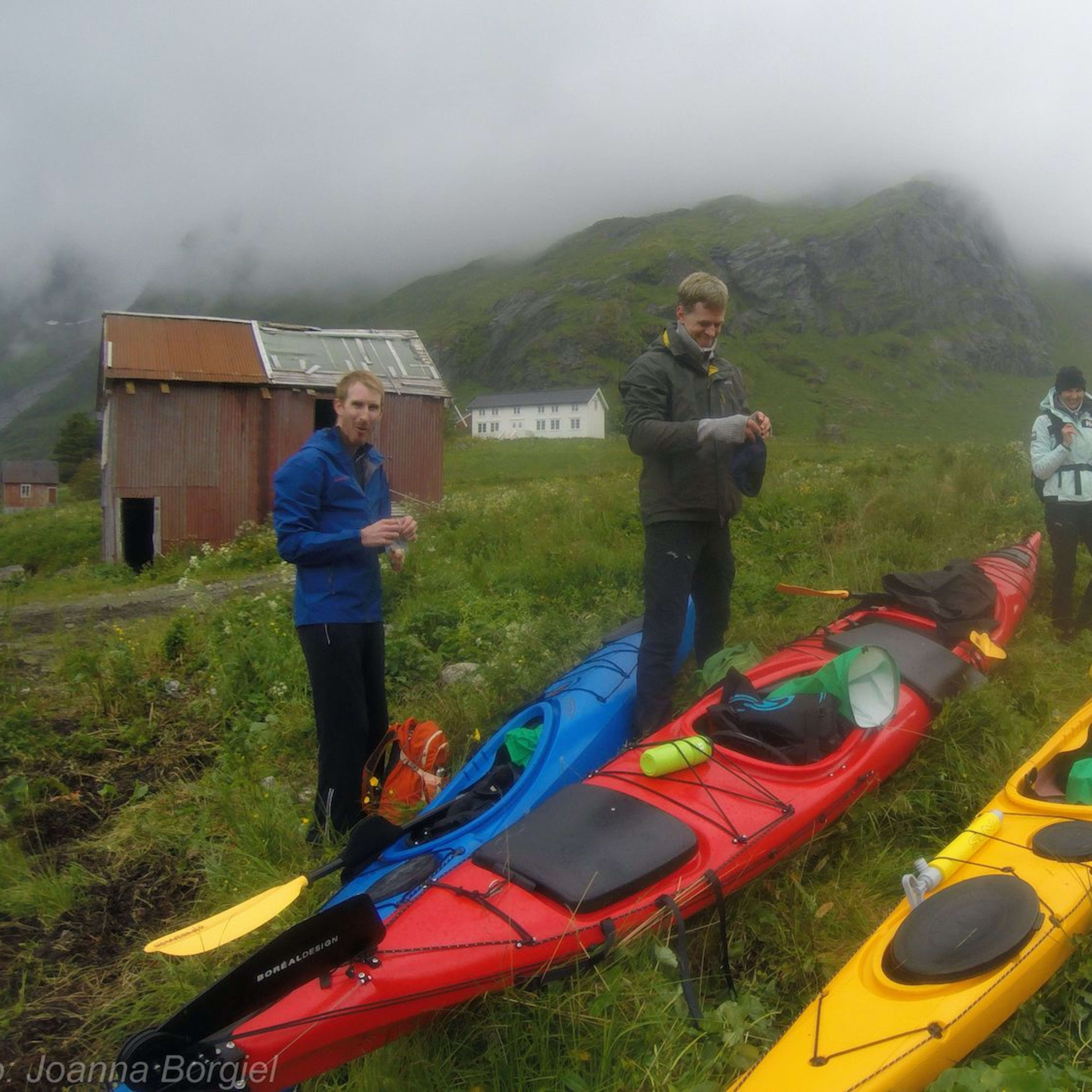 Group of kayakers preparing colorful kayaks on a grassy field with misty mountains in the background and rustic buildings.