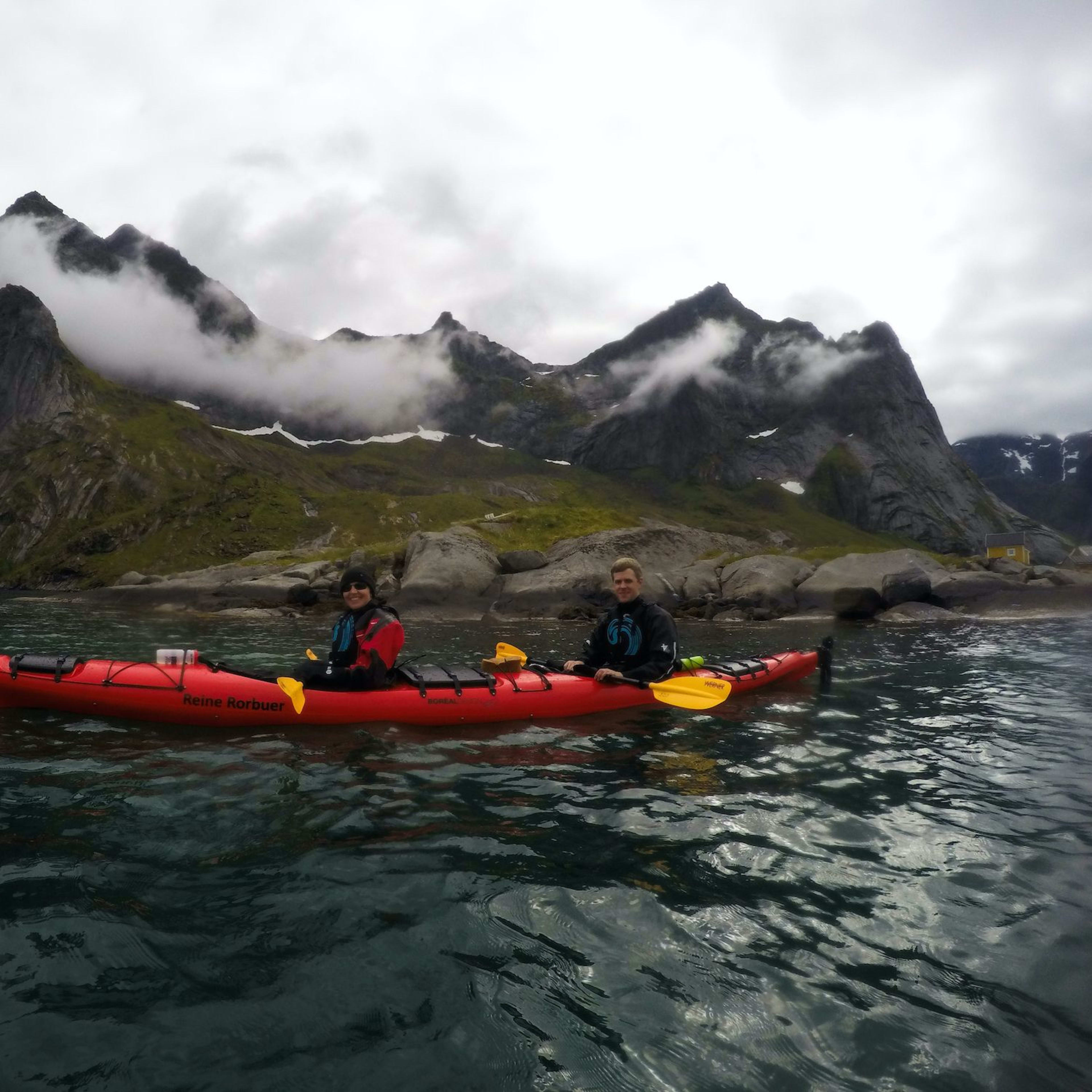 Two kayakers in a red tandem kayak labeled "Reine Rorbuer," paddling on calm, clear waters with green mountains and misty peaks.