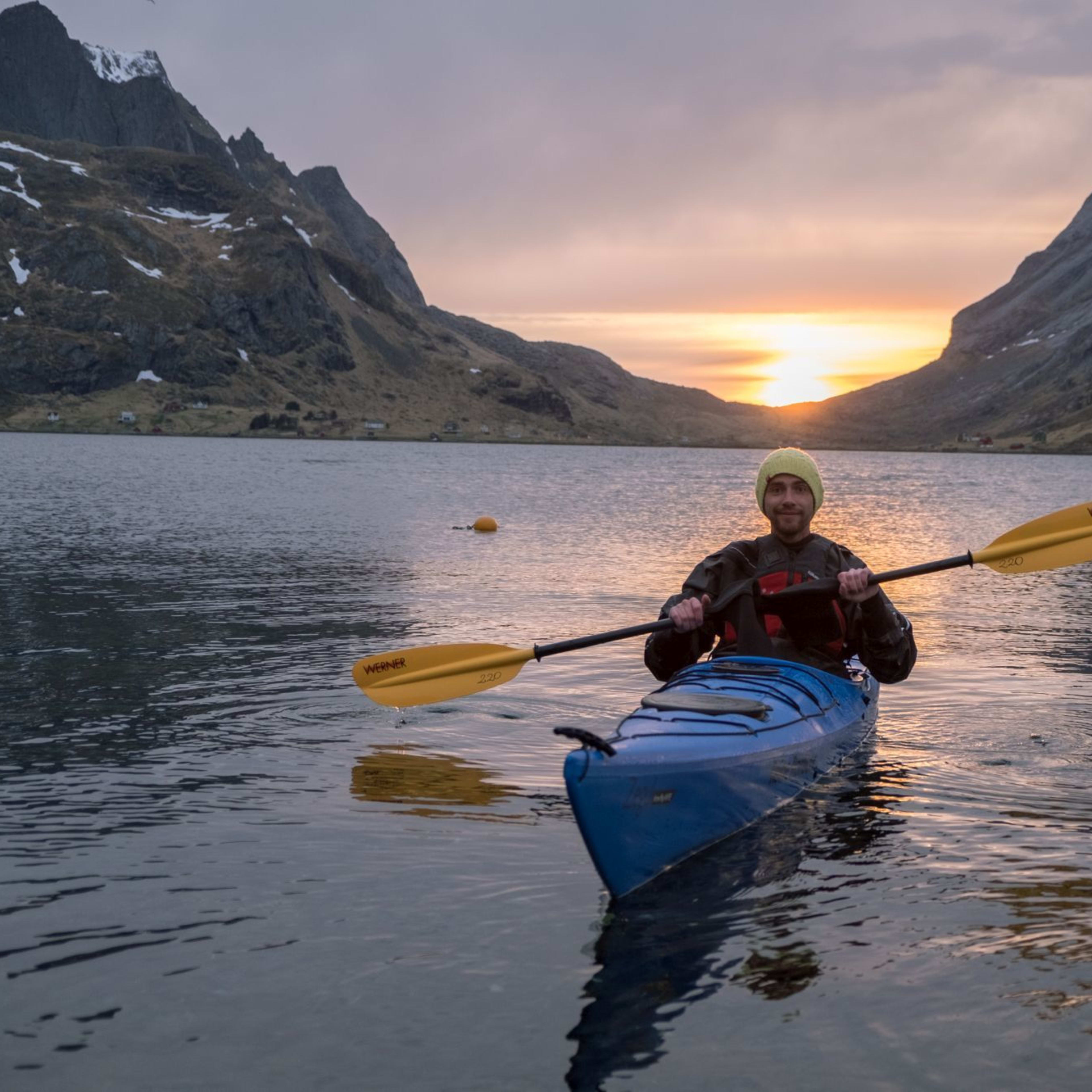 En person som padler på en lys blå kajakk med gule padler ved solnedgang, med gyldne refleksjoner på vannet og dramatiske fjell i bakgrunnen.
