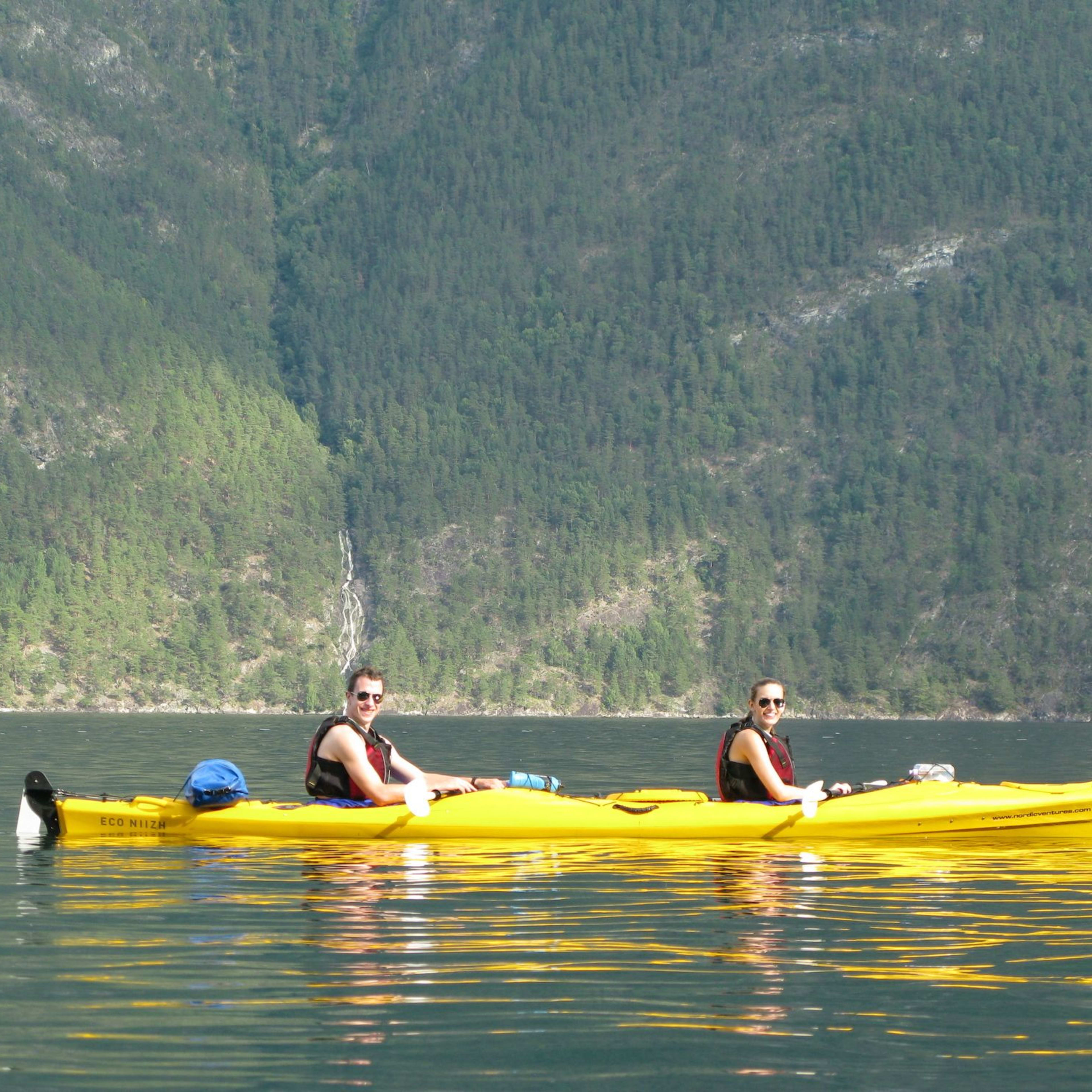 Two kayakers in a yellow tandem kayak on calm water, surrounded by a lush green forest and a narrow waterfall cascading down steep hills.