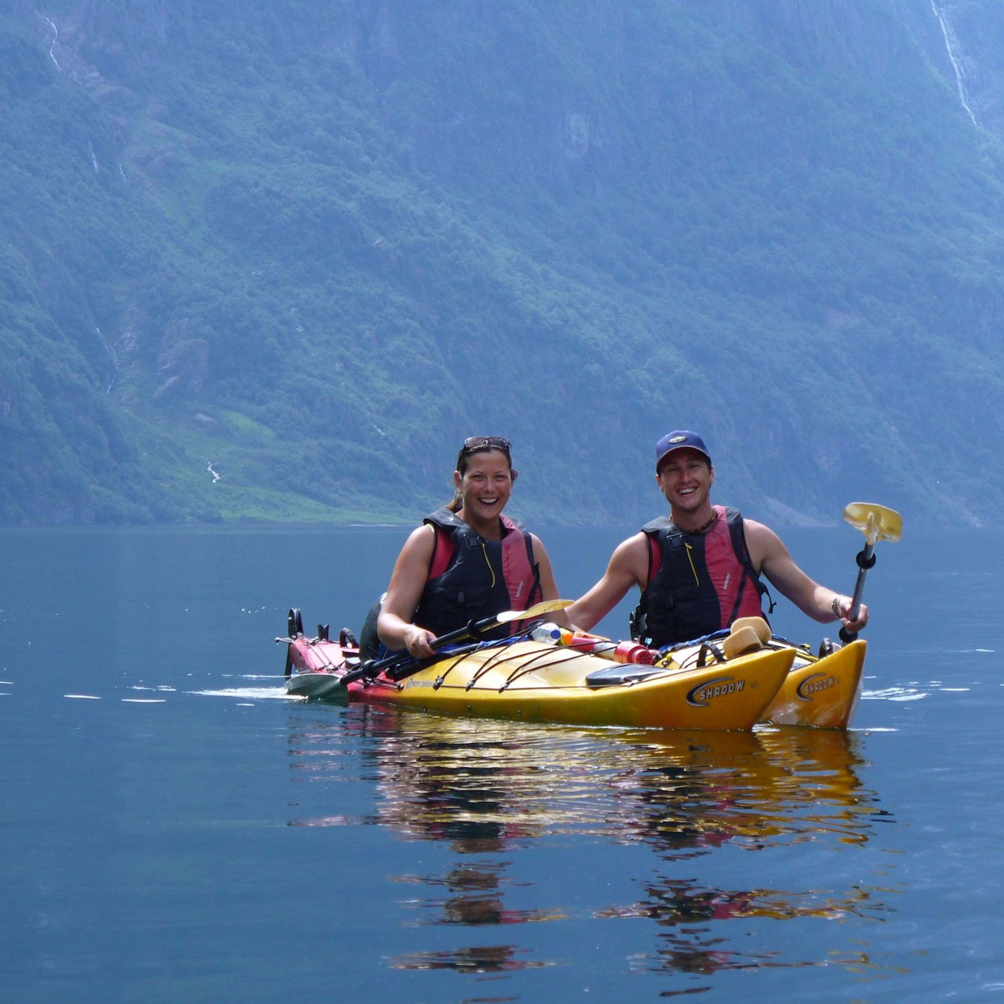 Two smiling kayakers in a yellow tandem kayak on calm blue water, surrounded by green hills and waterfalls in a peaceful fjord.