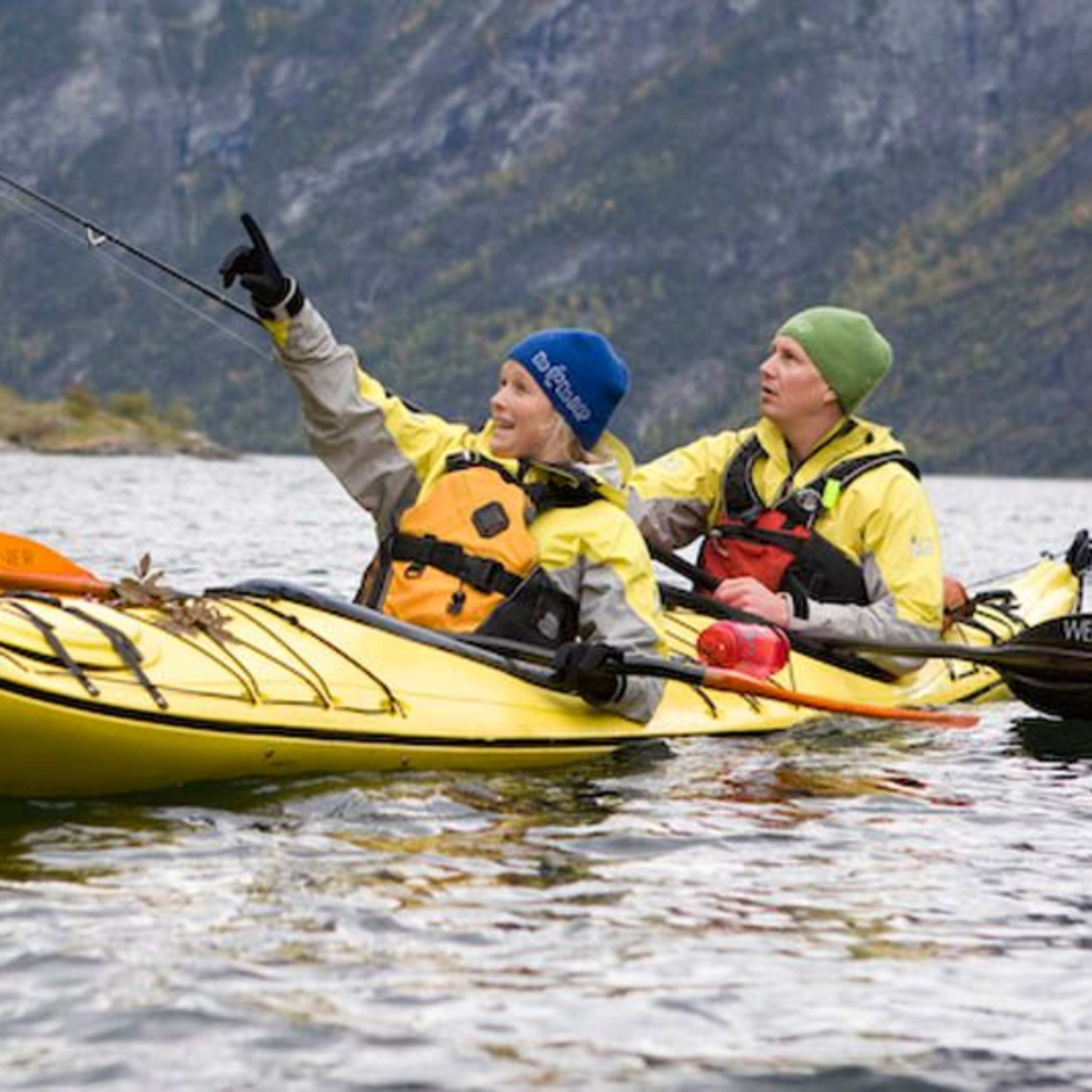 Zwei Kajakfahrer in einem gelben Tandemkajak paddeln auf einem ruhigen Fjord, wobei einer enthusiastisch auf etwas in der Ferne zeigt, umgeben von felsigen Klippen und Herbstlaub.