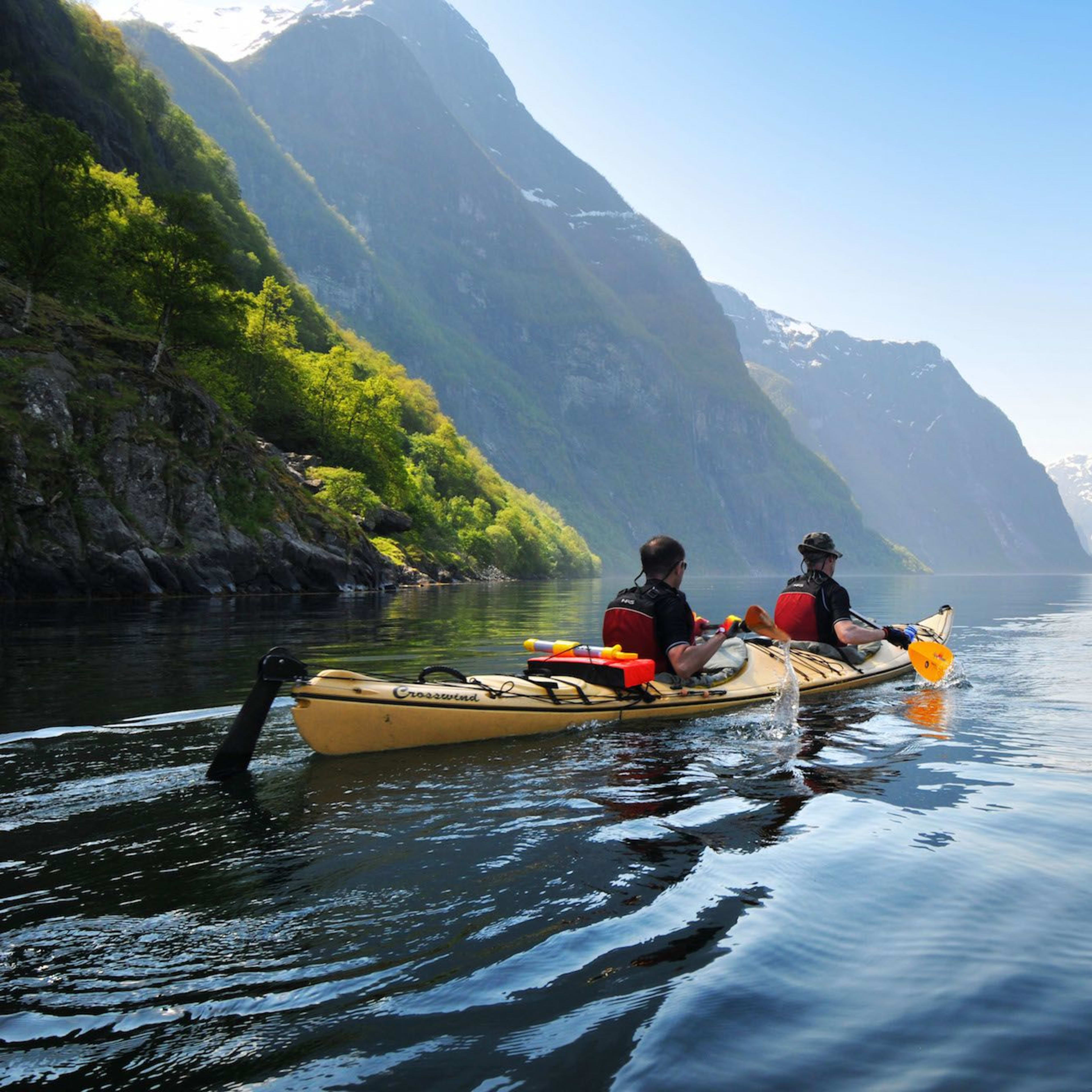 Two kayakers paddling along a calm, reflective fjord surrounded by towering green mountains and dramatic cliffs under clear blue skies.