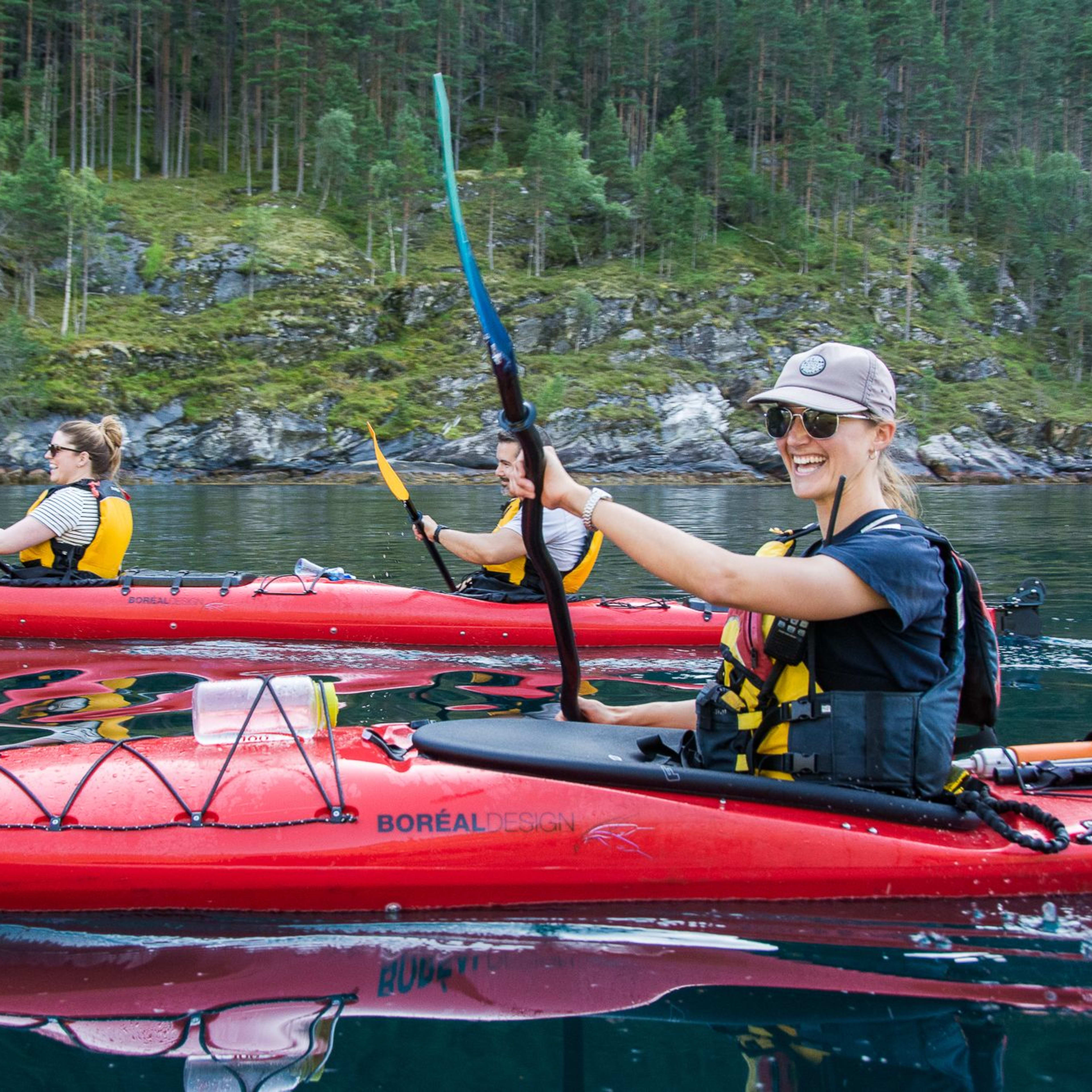 Woman kayaking in a red kayak, smiling and holding her paddle with one hand while paddling, with reflections of the landscape in calm water.