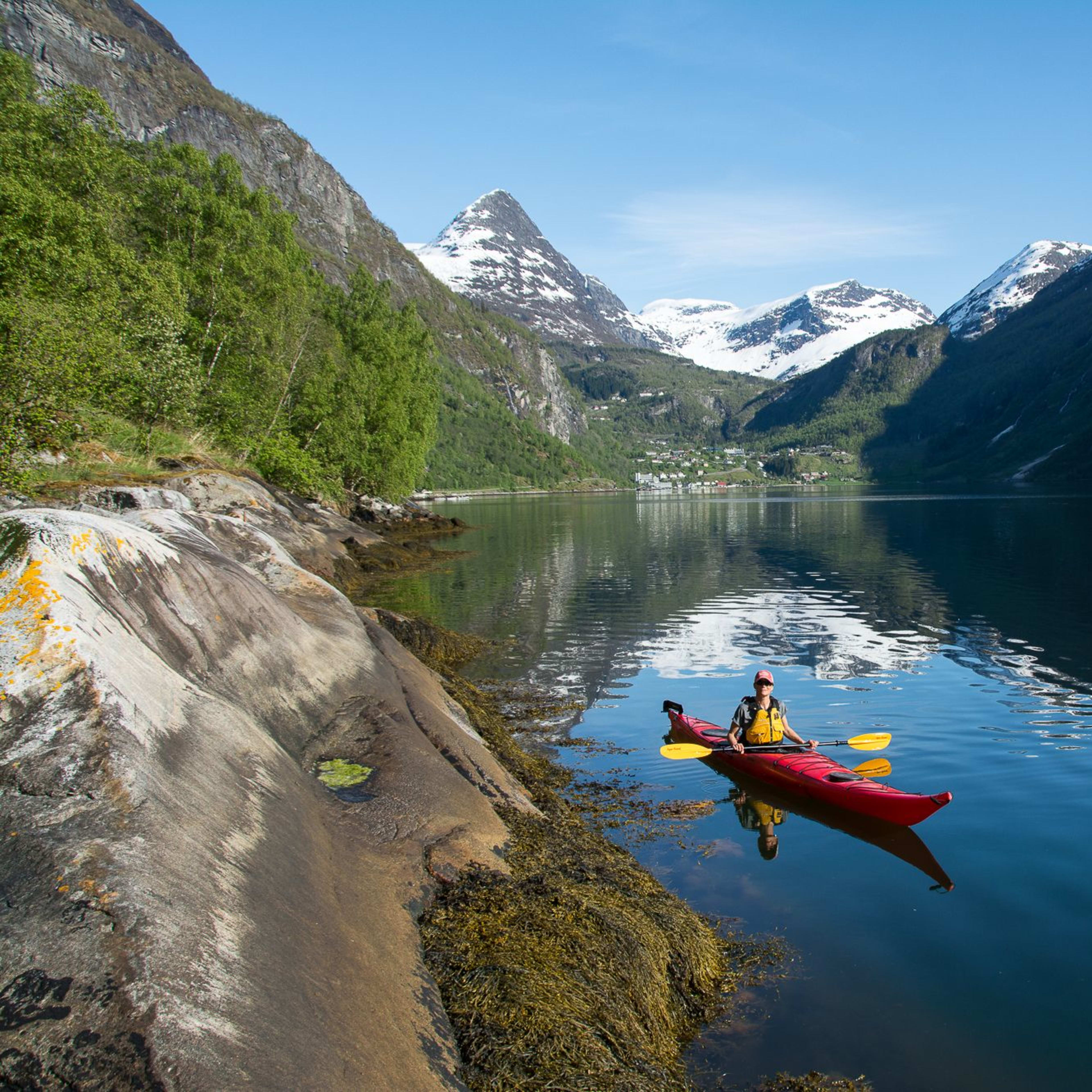 Person kayaking in a red kayak on calm waters, with snow-covered mountains and a small village visible in the background.