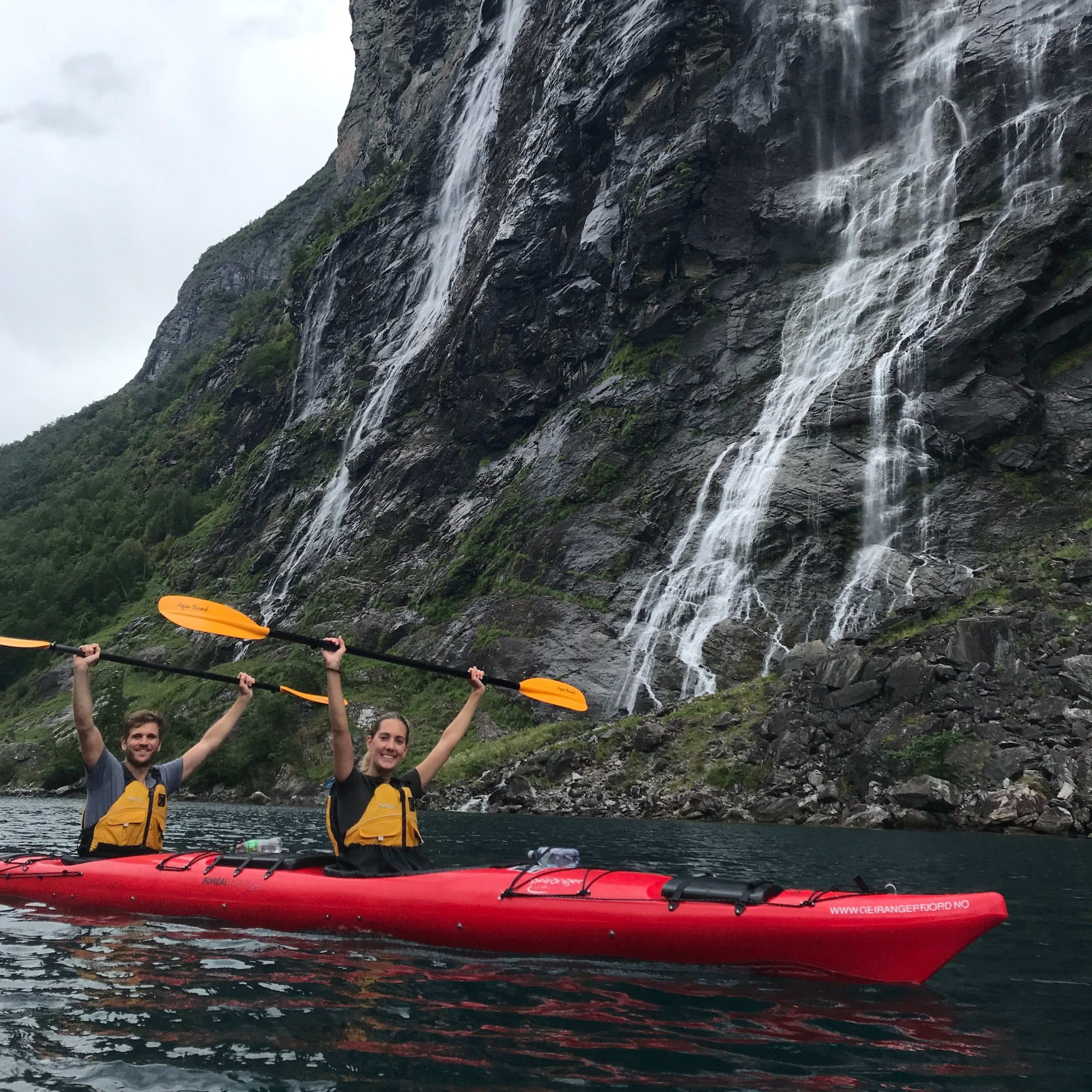 Man and woman kayaking in a red kayak on calm waters, raising paddles triumphantly, surrounded by cliffs and waterfalls.