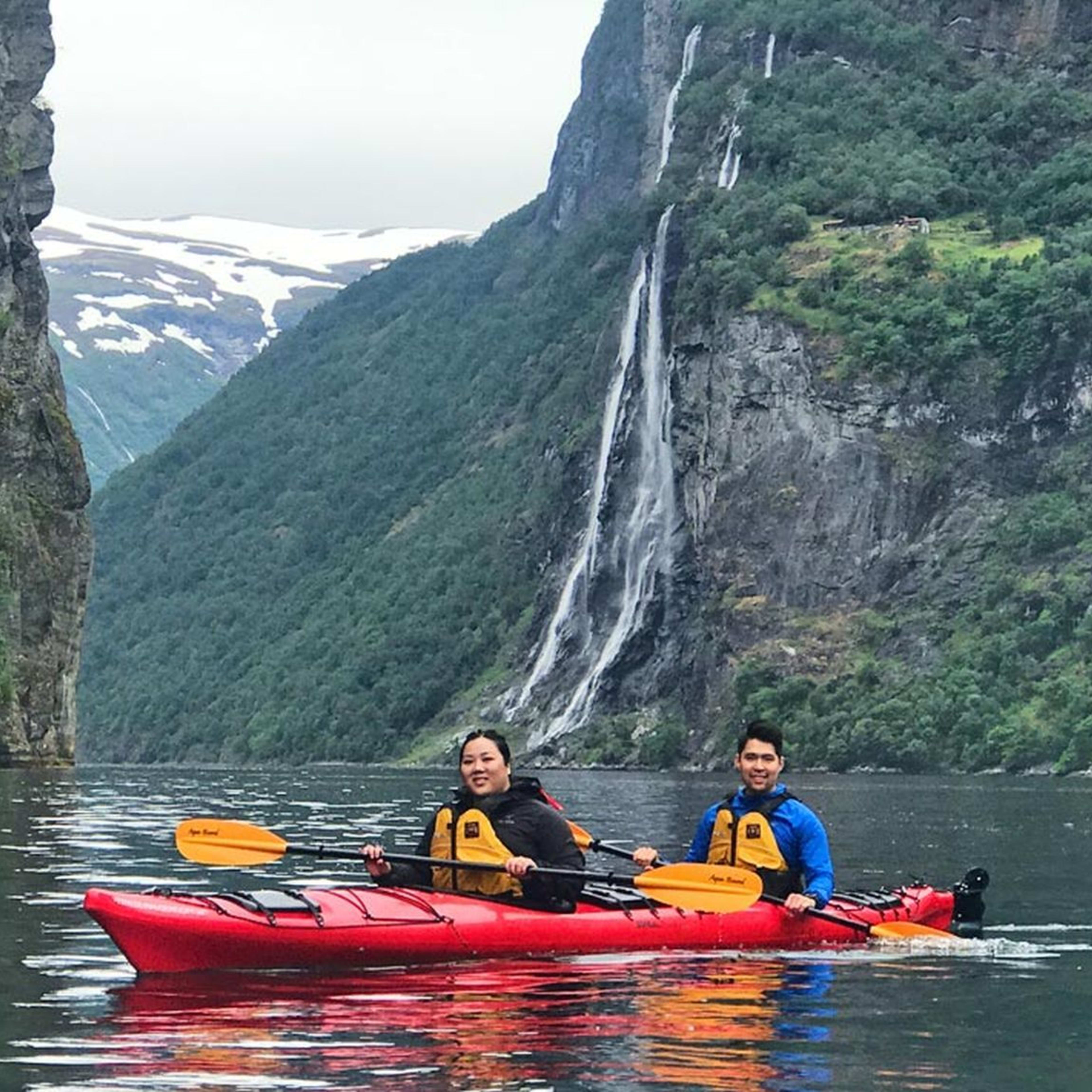 Zwei Kajakfahrer paddeln zusammen in einem roten Kajak durch einen norwegischen Fjord mit einem Wasserfall und schneebedeckten Bergen.