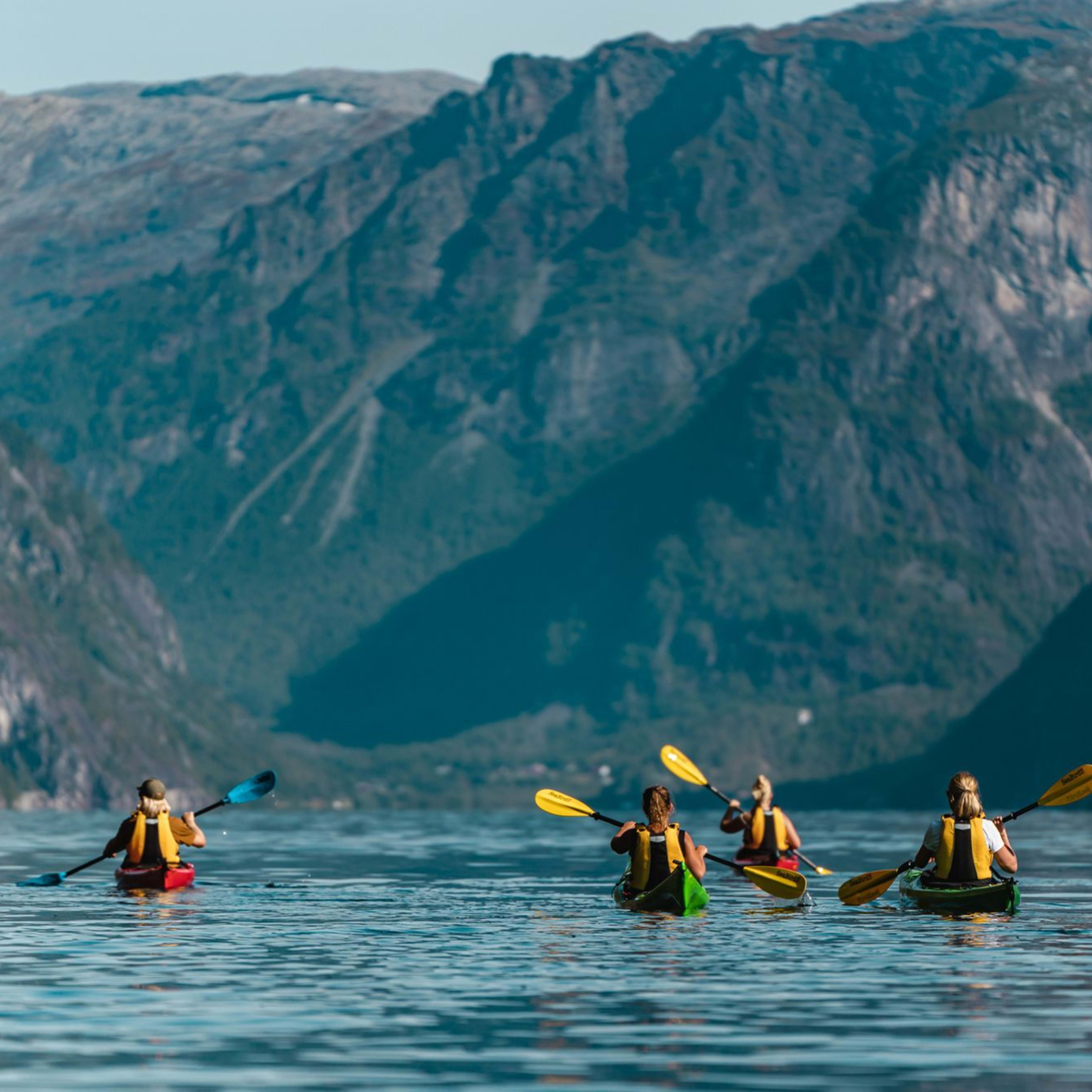 Group of four kayakers paddling on calm water with dramatic mountains and colorful kayaks, reflecting on the peaceful water.
