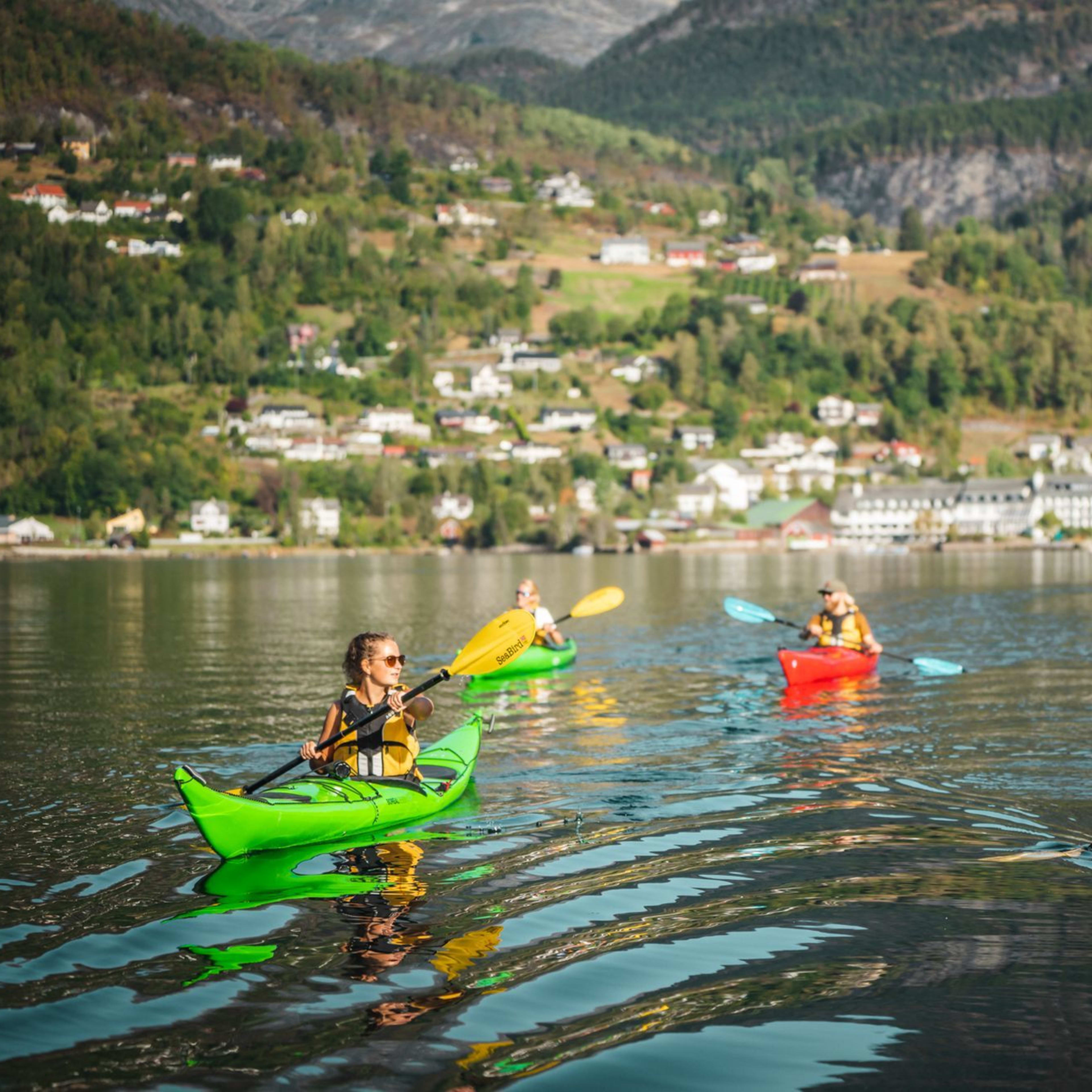 Eine Gruppe von Kajakfahrern auf ruhigem Wasser mit einem Dorf im Hintergrund, paddelnd in einer Linie mit grünen, gelben und roten Kajaks unter einem klaren Himmel.