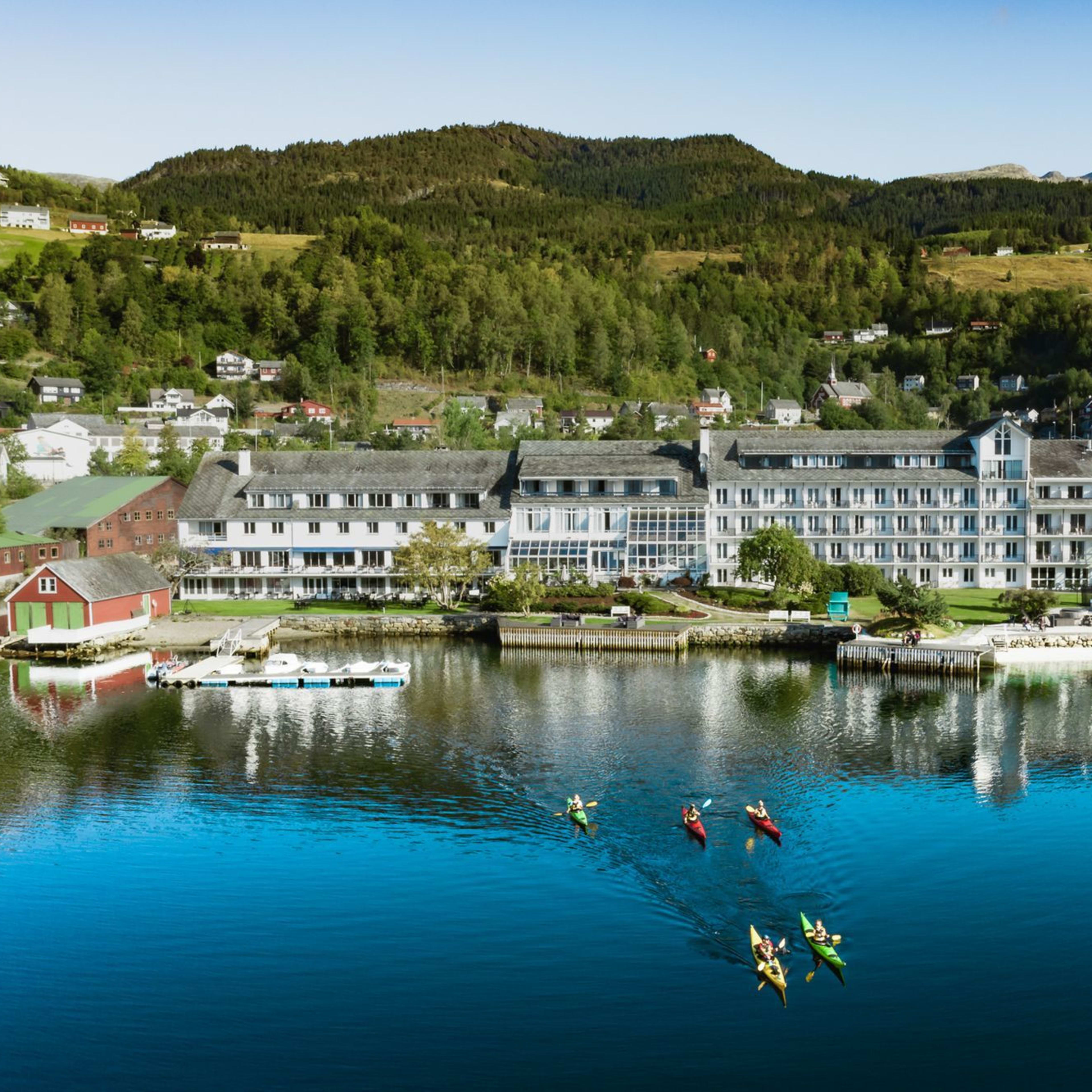 Eine Gruppe von Kajakfahrern paddelt vor einem großen Hotel mit einem langen weißen Gebäude, umgeben von grünen Hügeln, mit Reflexionen im ruhigen Wasser.