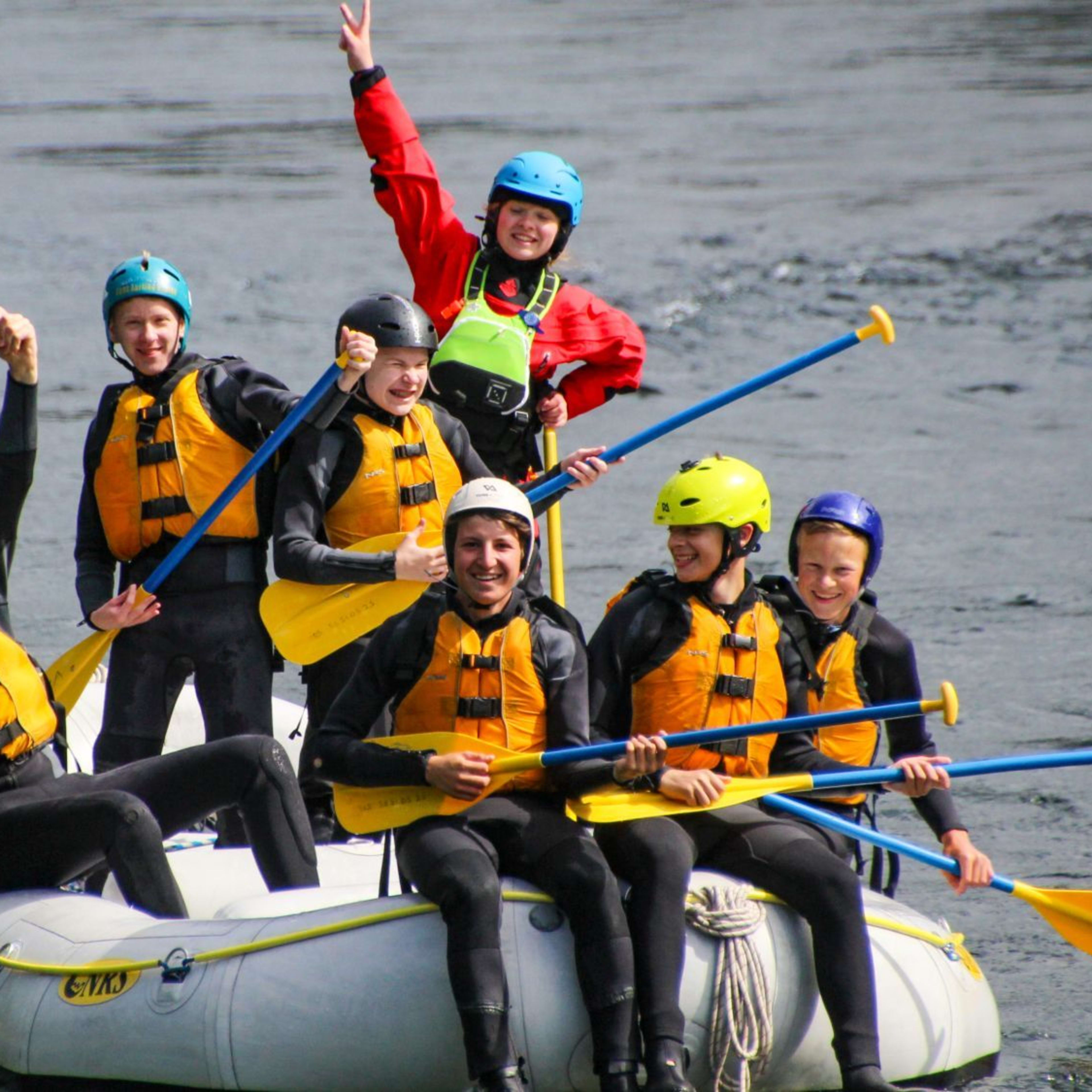 A group rafting joyfully on a river in a white raft, with bright-colored helmets and life jackets, paddling through calm water with ripples.