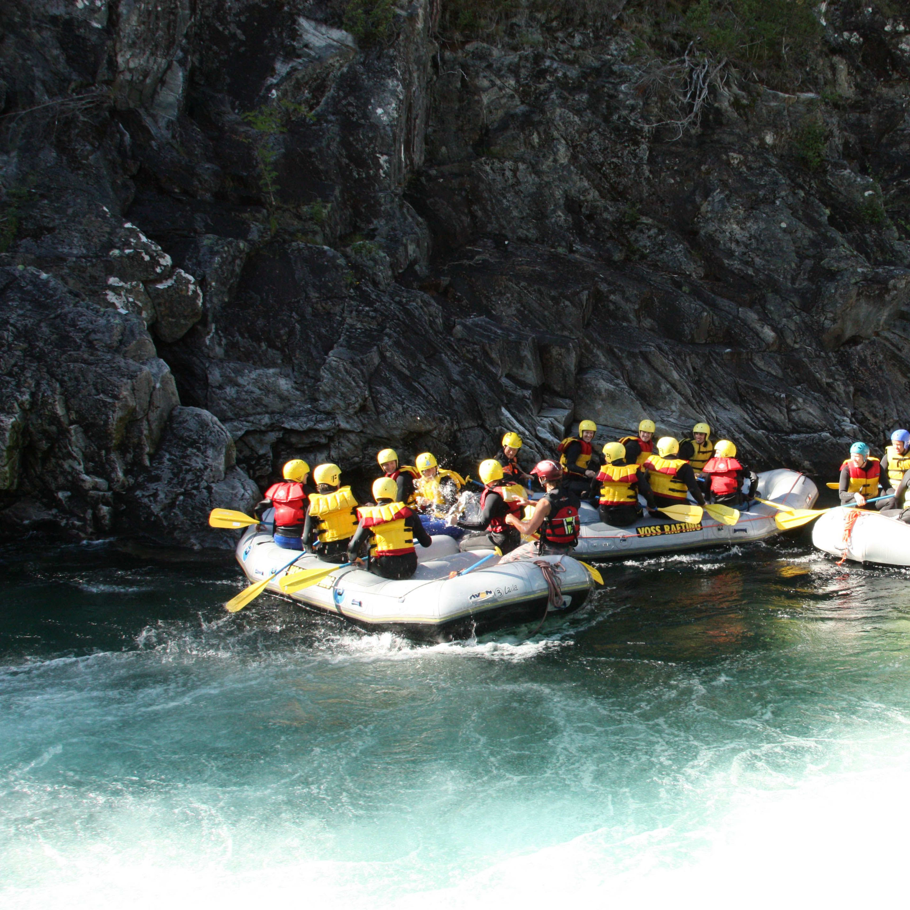 Mehrere Gruppen von Menschen paddeln in der Nähe von felsigen Klippen, einige interagieren mit Guides am Flussufer.