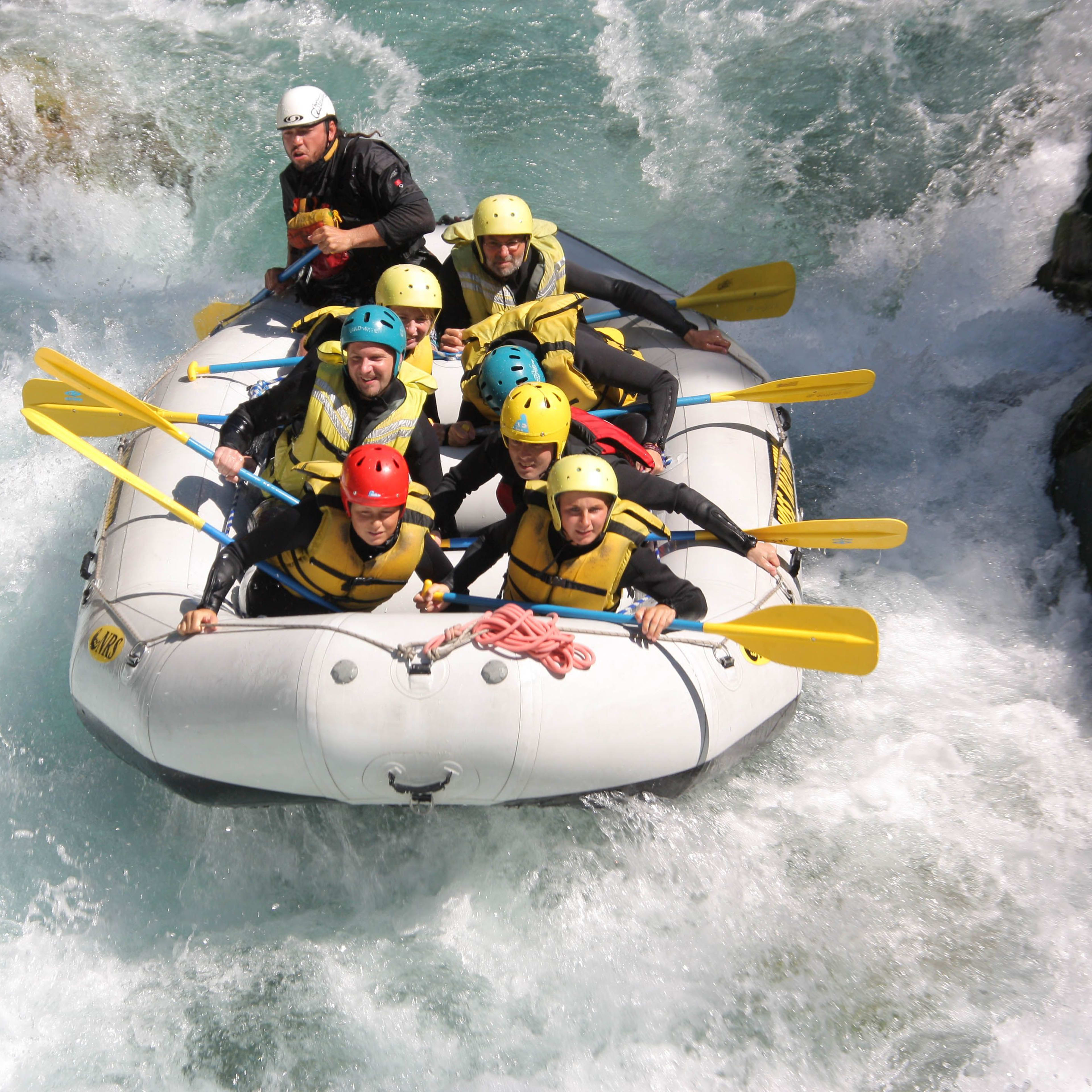 Eine Gruppe von Menschen navigiert durch Stromschnellen in einem Raft, mit felsigem Gelände und üppigem Grün um sie herum.
