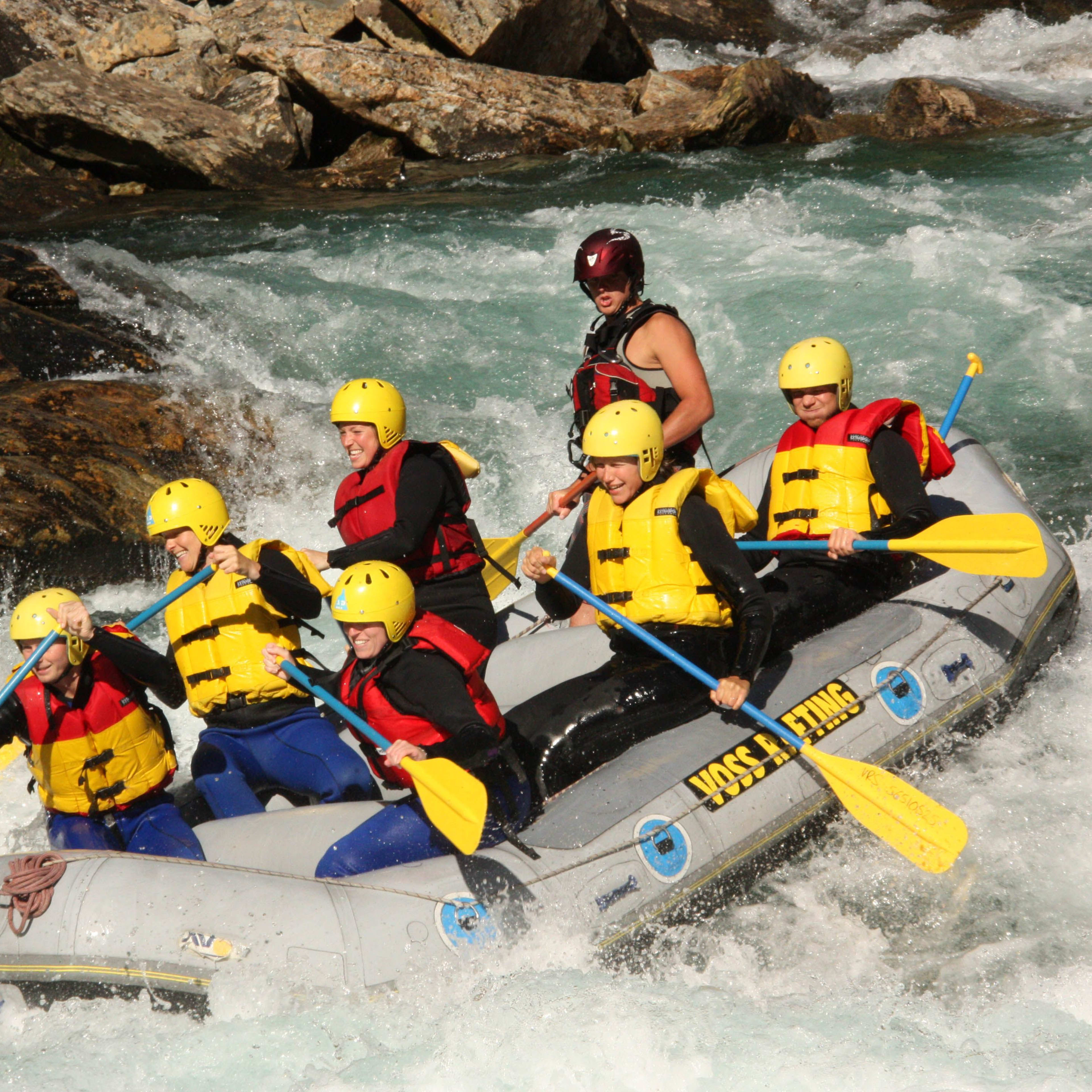 Sieben Personen paddeln in einem Raft durch schnell fließendes Wasser, mit einem Guide, der die Richtung steuert.