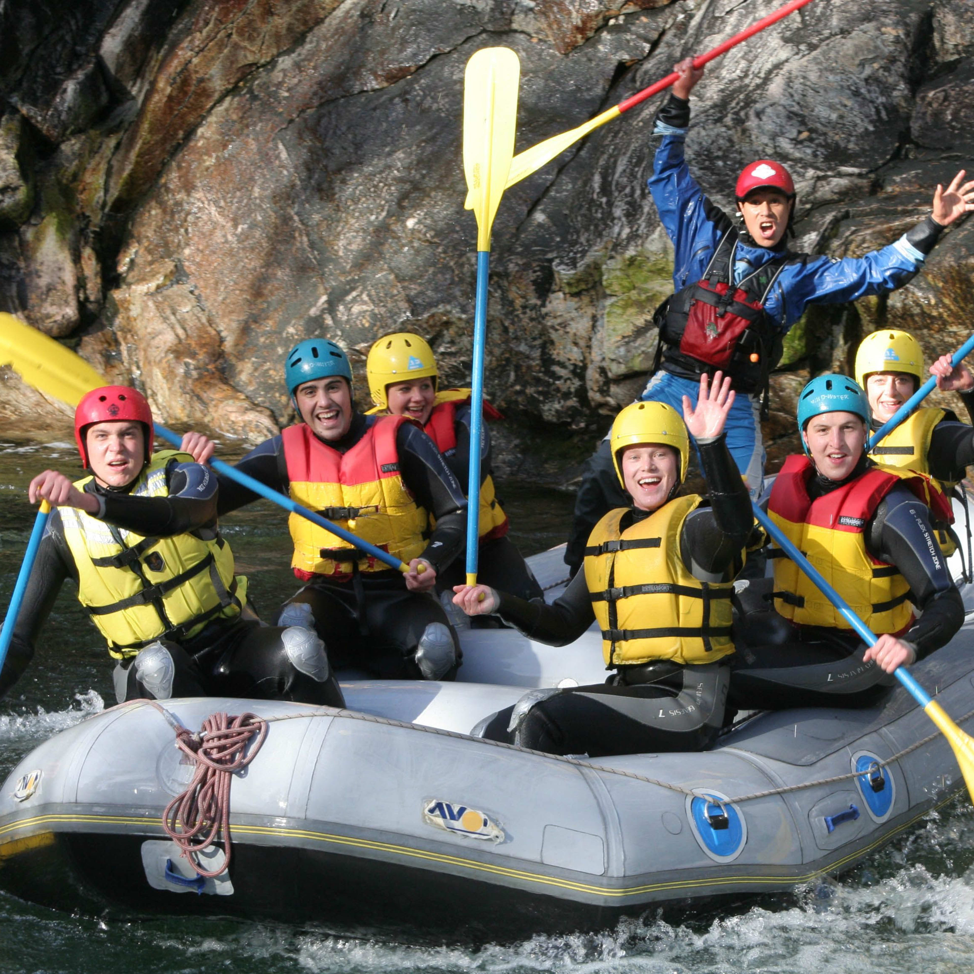 Acht Personen in einem Raft, die lächeln und durch ruhiges Wasser paddeln, mit einem Flussführer im Hintergrund.