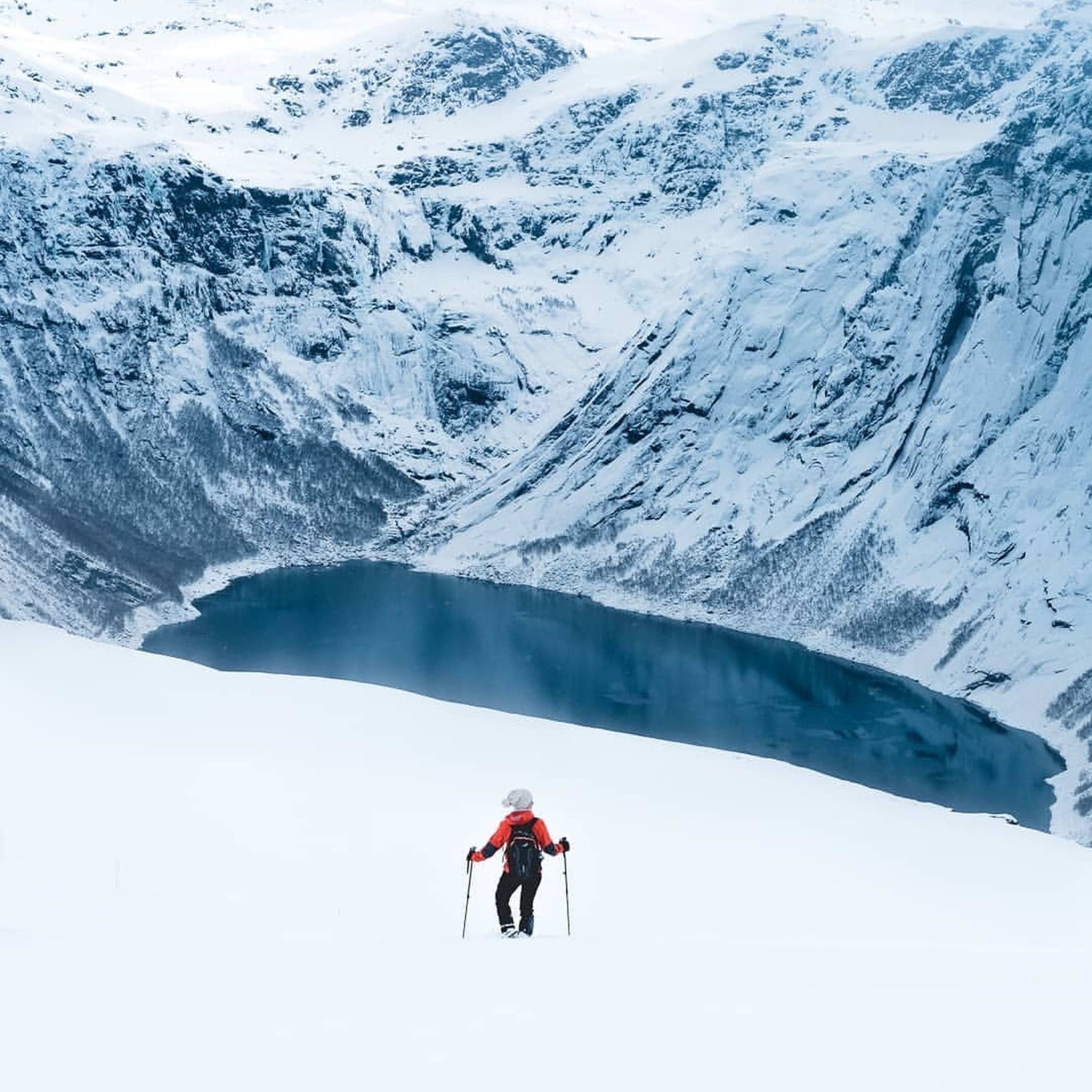 Ein einsamer Wanderer in roter Jacke und Schneeschuhen wandert über eine verschneite Landschaft und nähert sich einem Fjord, der von schneebedeckten Bergen umgeben ist.
