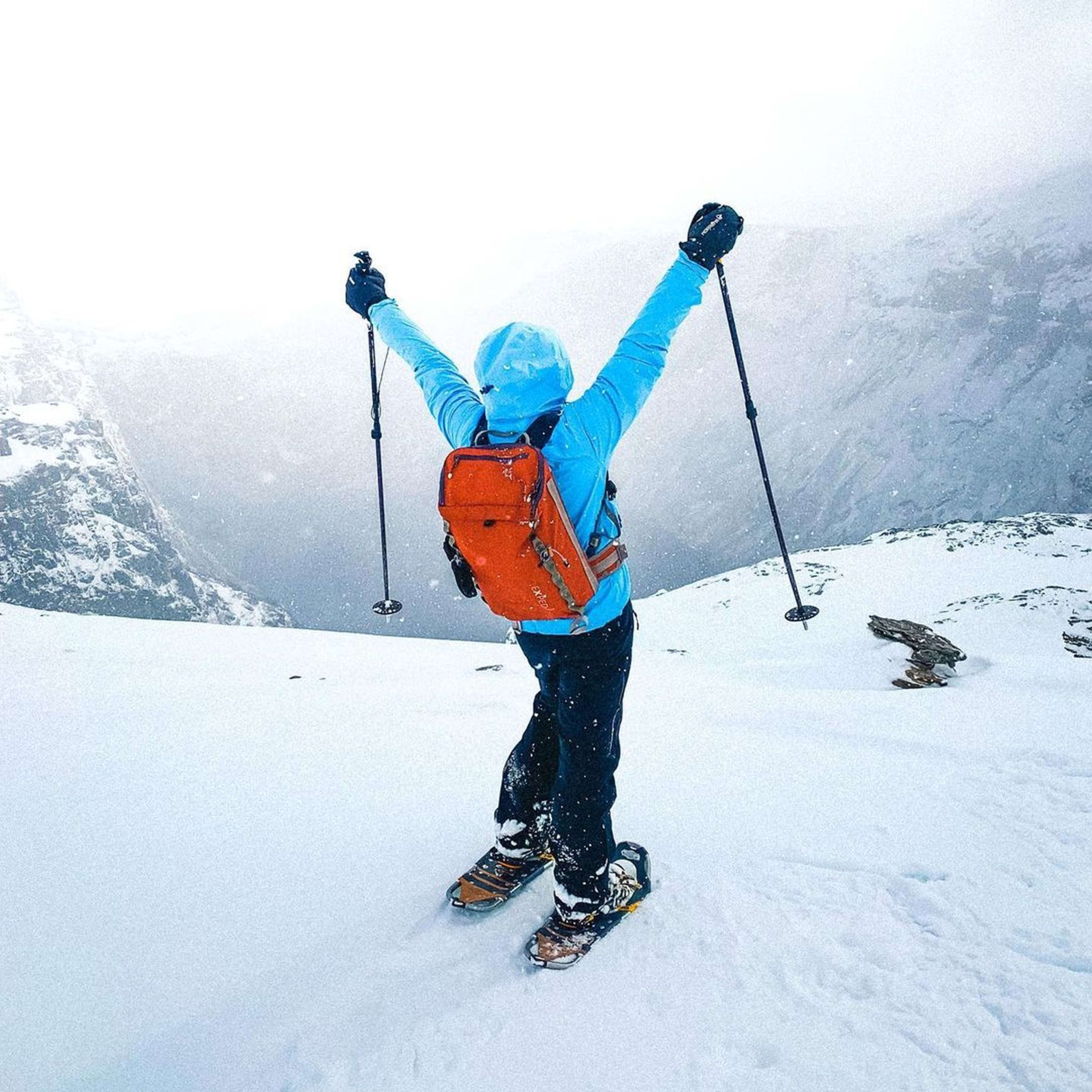 Eine Person in blauer Jacke und orangefarbigem Rucksack steht triumphierend im tiefen Schnee mit Wanderstöcken auf einem schneebedeckten Bergrücken.
