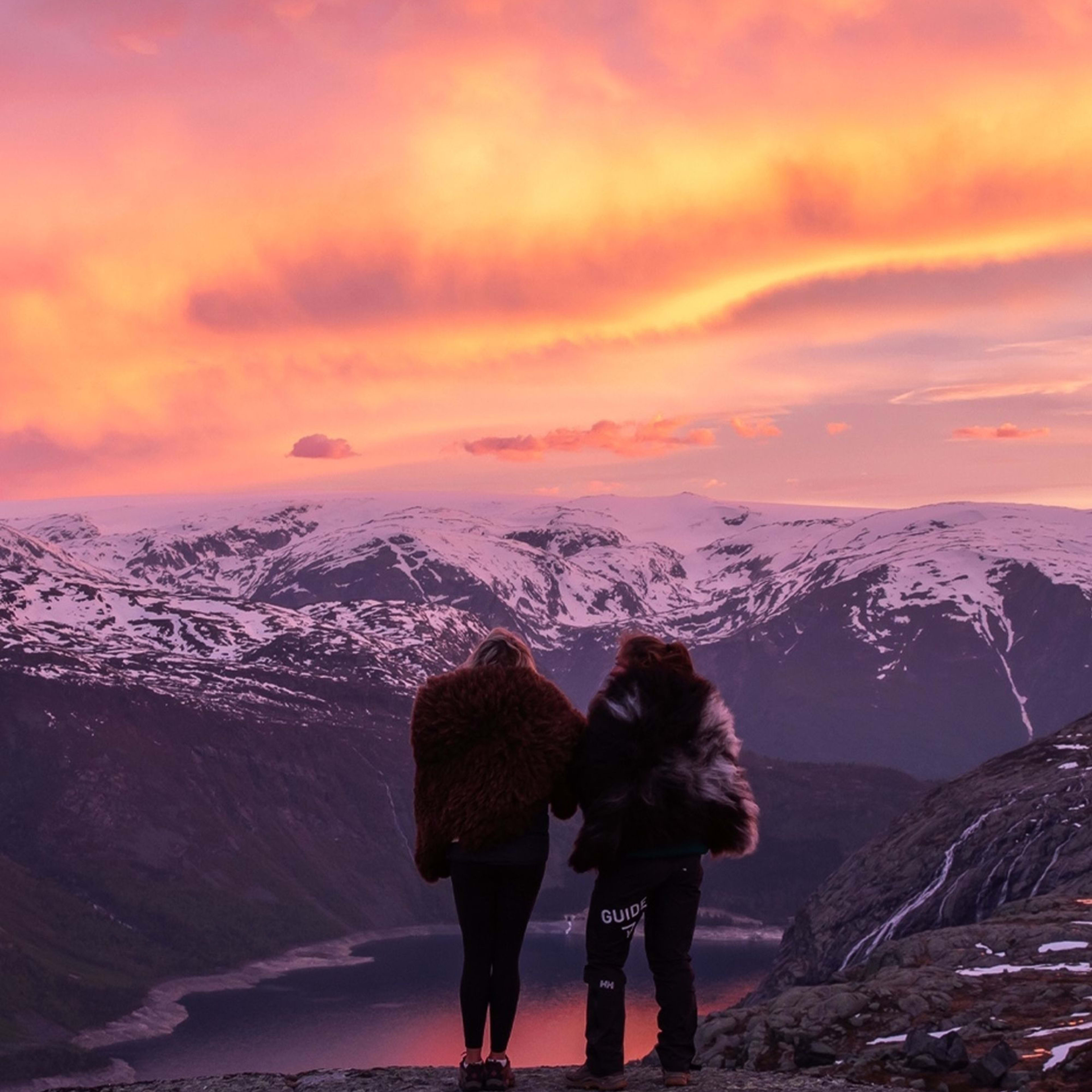 Zwei Personen in Fellmänteln stehen auf einem Bergrücken und beobachten einen Sonnenuntergang über schneebedeckten Gipfeln und einem Fjord.