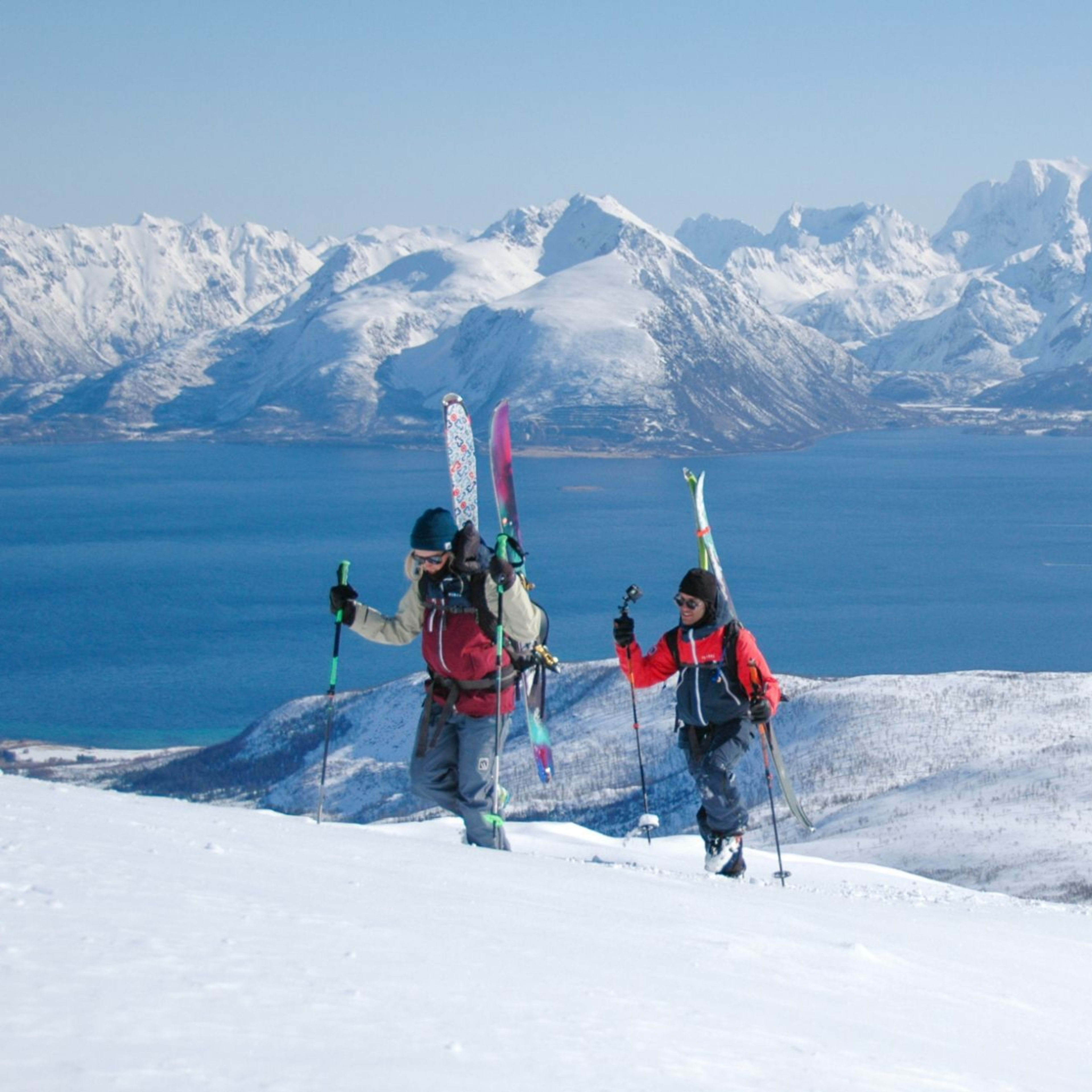Two skiers hike uphill through snow with skis on their backpacks, surrounded by snowy mountains and a deep blue fjord.