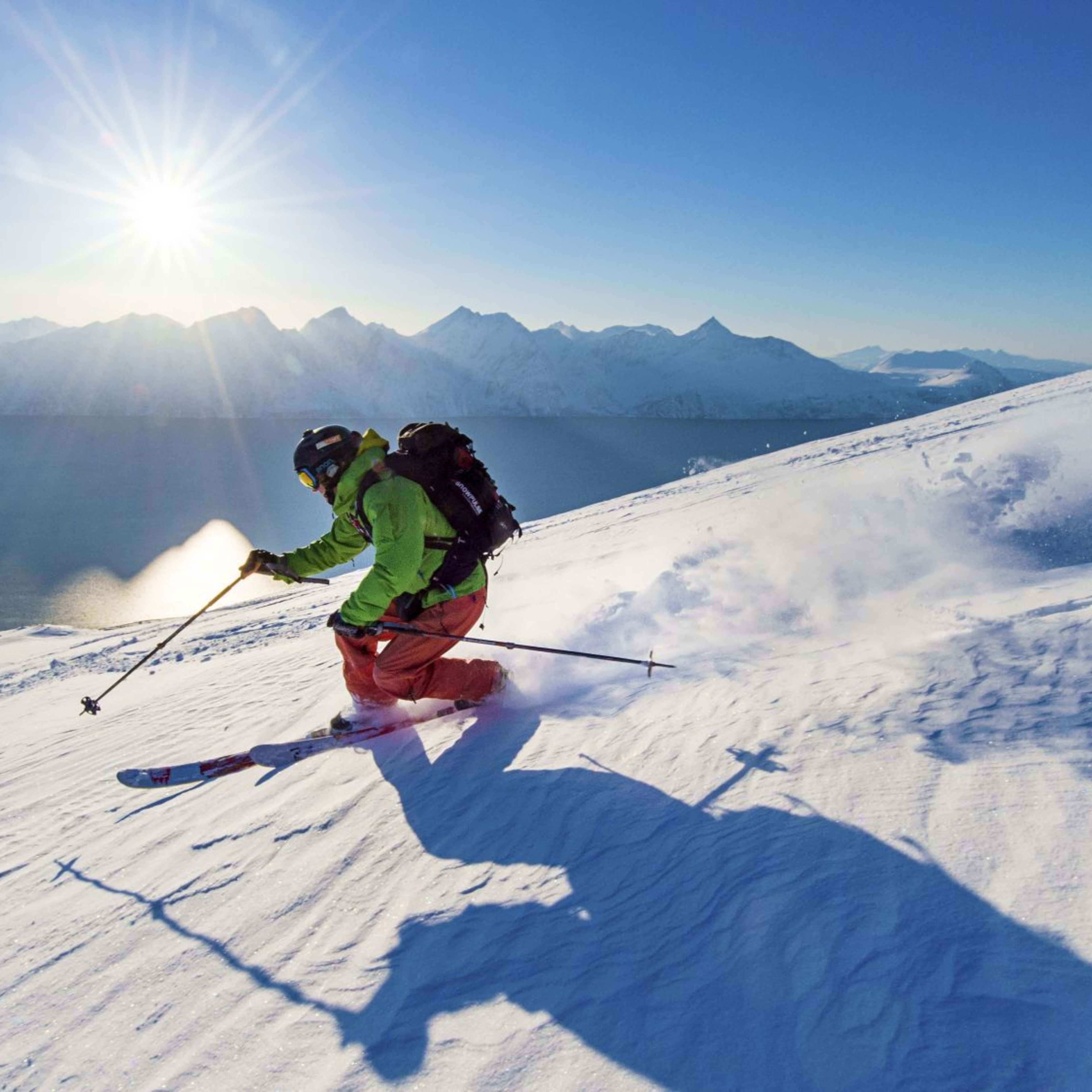 A skier in bright gear glides down a sunlit snowy slope, casting a long shadow, with a calm fjord and rugged mountains in the background.