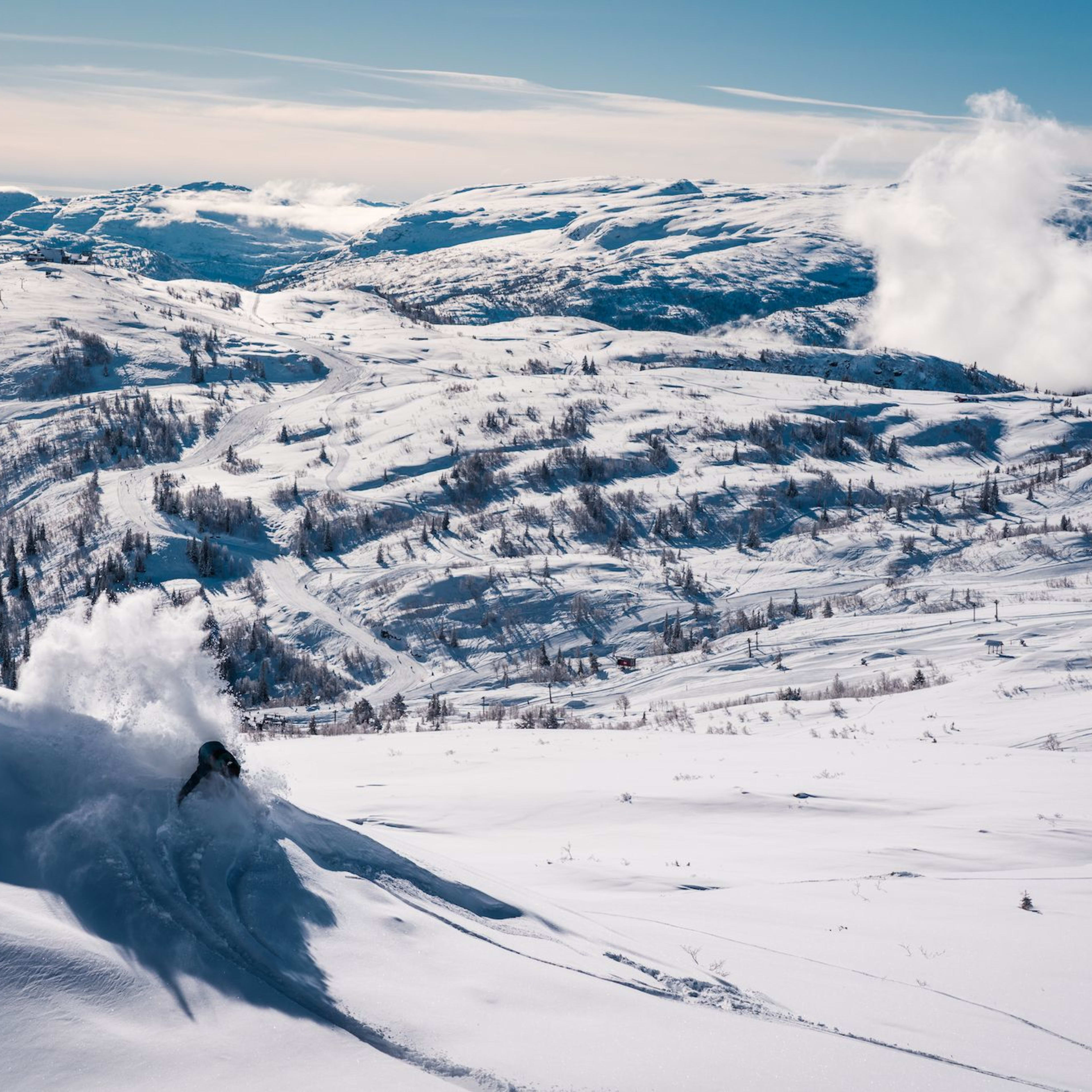 En skientusiast skjærer ned en bratt snødekt bakke og etterlater et spor av puddersnø, med fjell i bakgrunnen.