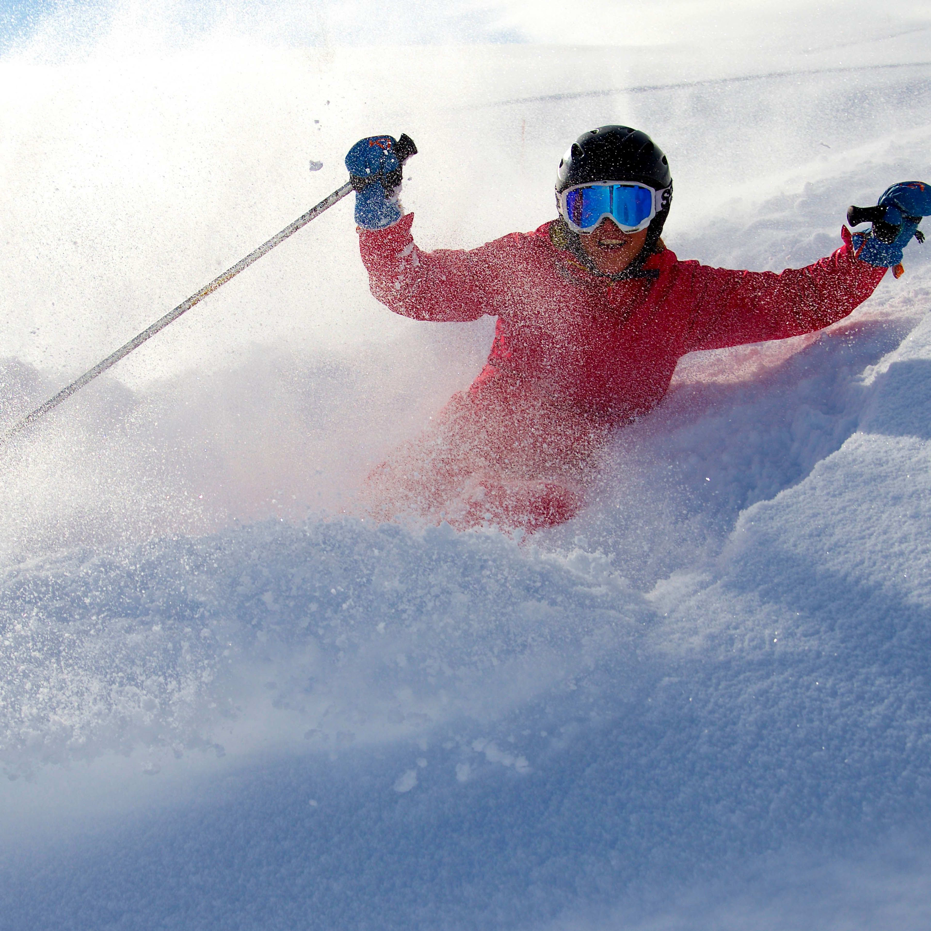 A skier in a red jacket and blue goggles plows through deep powder snow, creating a dramatic spray as they carve through a sunlit slope.