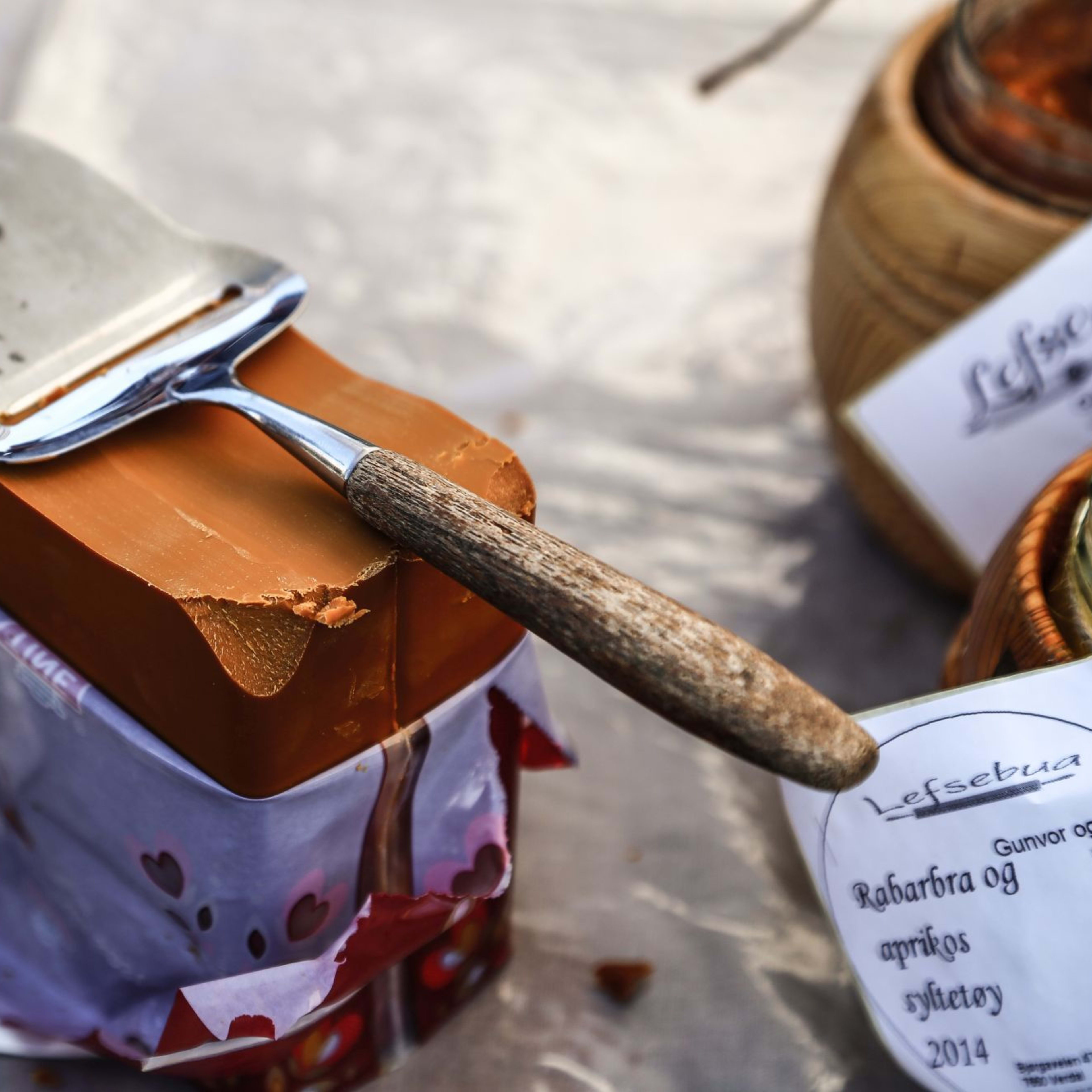 A block of brown Norwegian goat cheese sits on a plate with a cheese slicer resting on top, surrounded by jars and traditional food packaging.