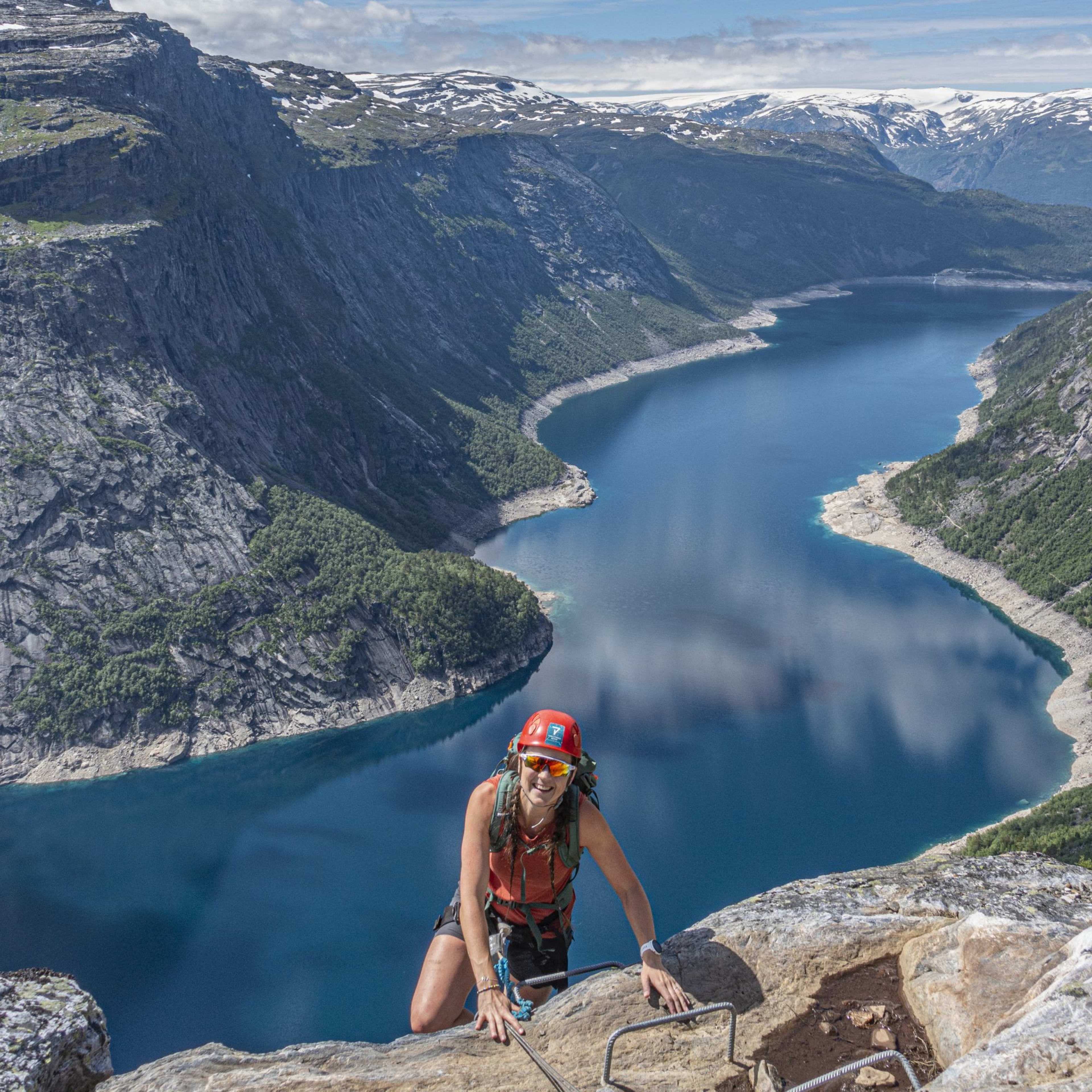 Climber in helmet and sunglasses smiles while scaling cliff over fjord.