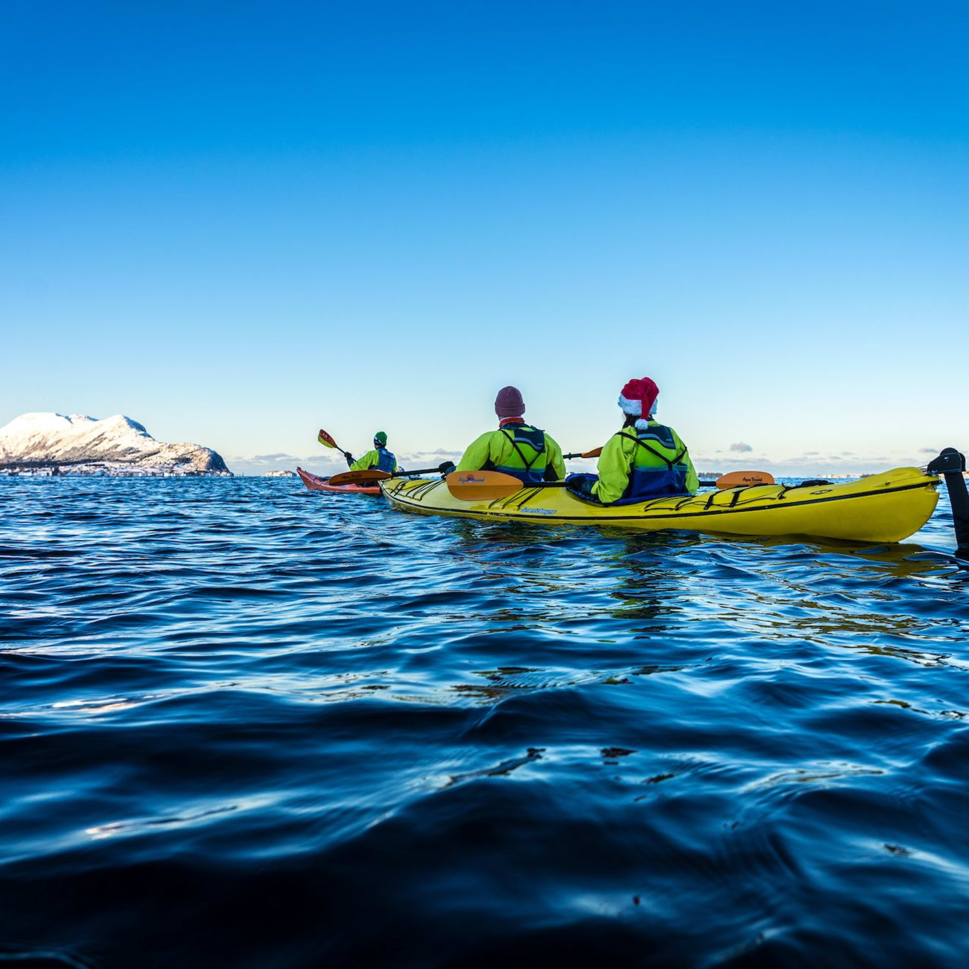 Drei Personen paddeln, wobei eine einen Weihnachtsmann-Hut trägt, auf dem Wasser mit schneebedeckten Bergen und klarem blauen Himmel im Hintergrund.