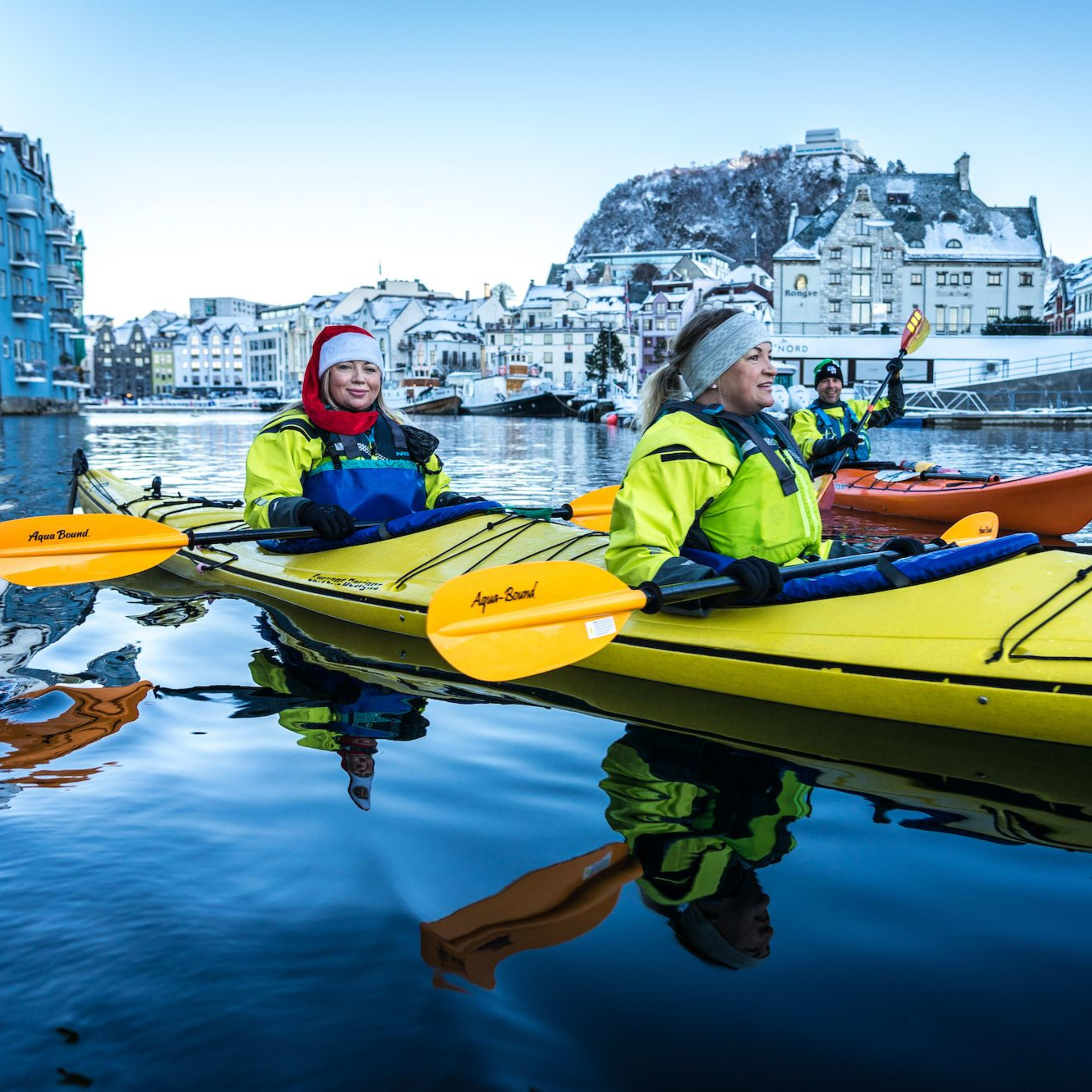 Drei Personen paddeln im Wasser, umgeben von schneebedeckten Gebäuden, mit leuchtend gelben und blauen Schwimmwesten.