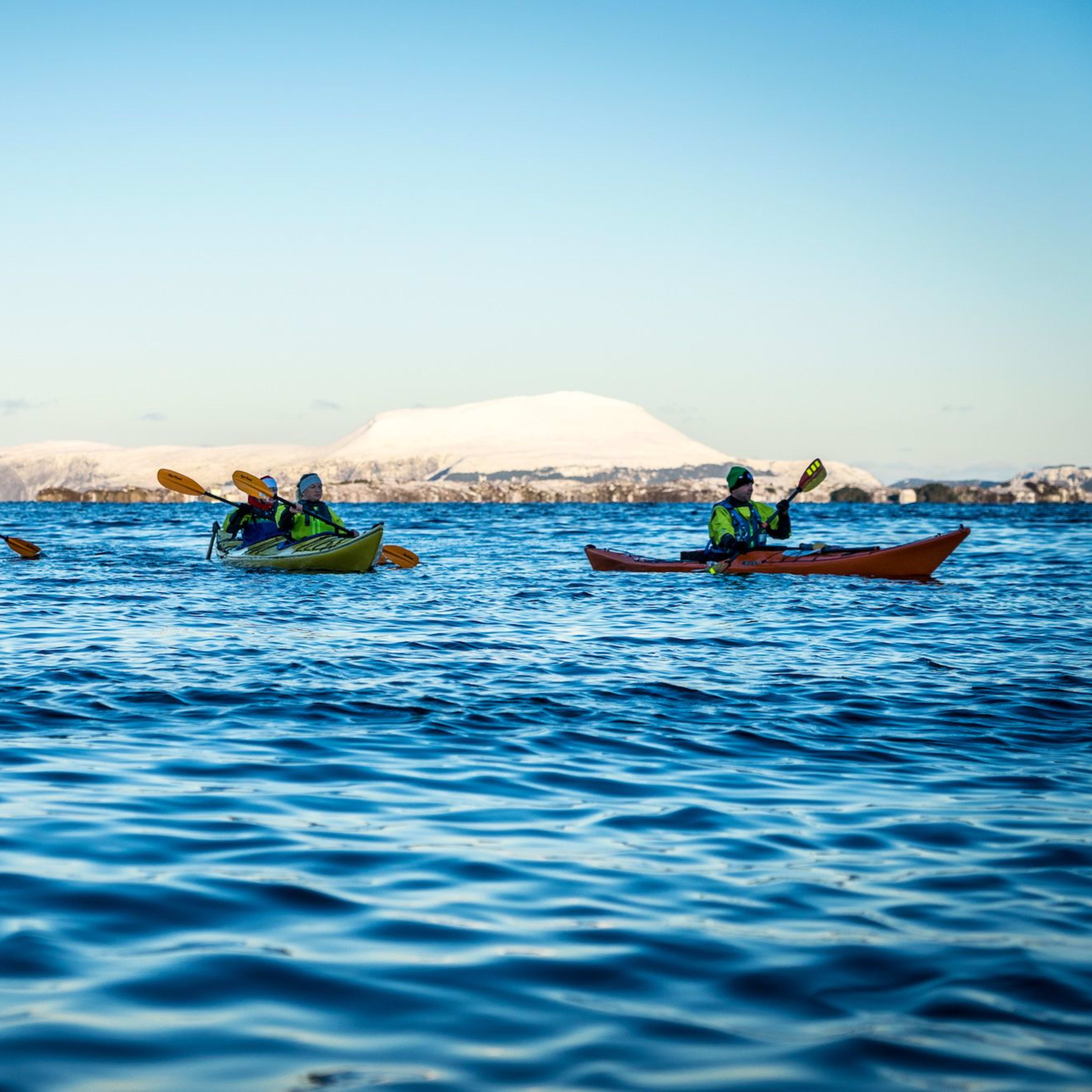 Fünf Personen paddeln auf ruhigem blauen Wasser, umgeben von schneebedeckten Bergen und Gebäuden entlang des Ufers.