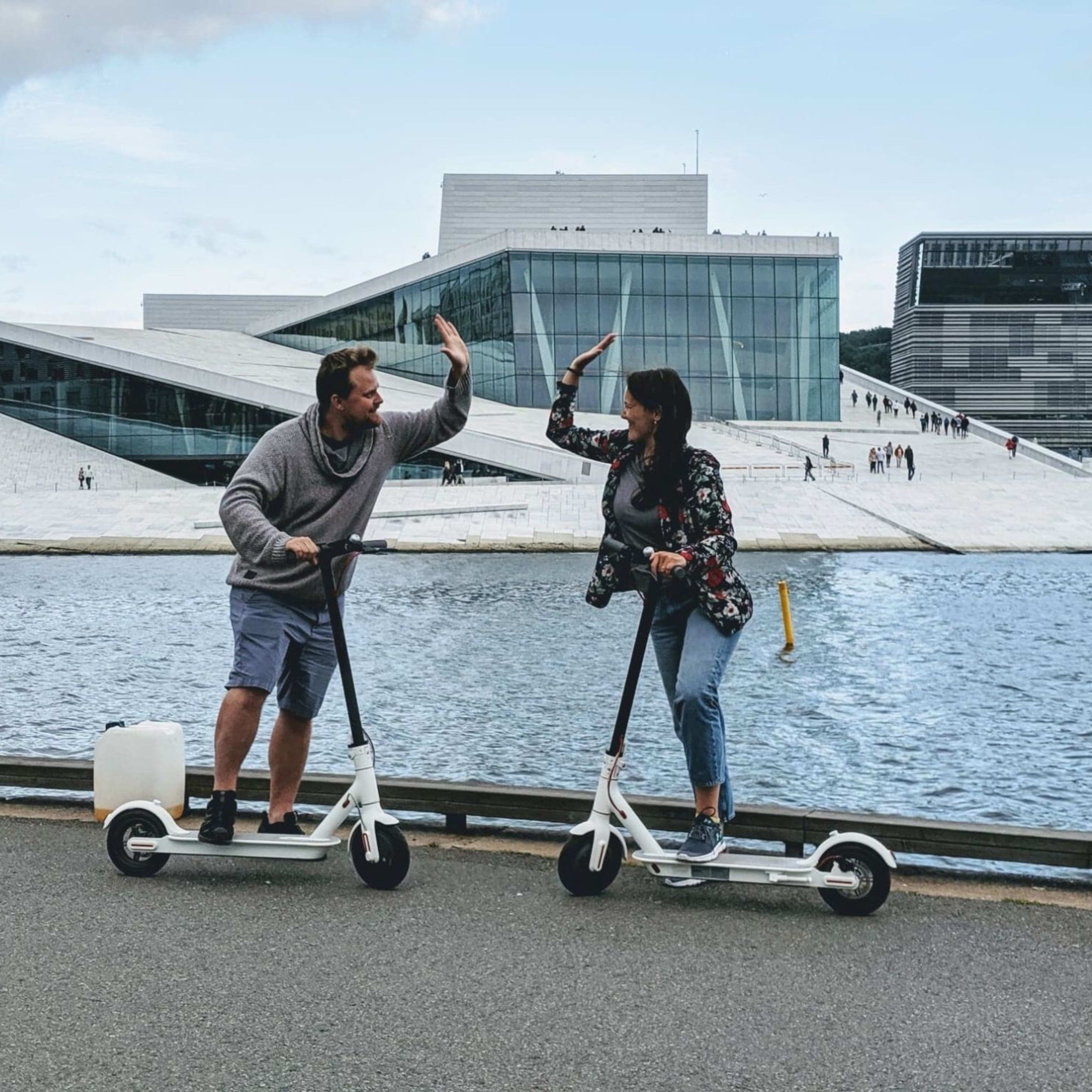 Mann und Frau auf E-Scootern geben sich High-Five vor der Oper in Oslo am Wasser.