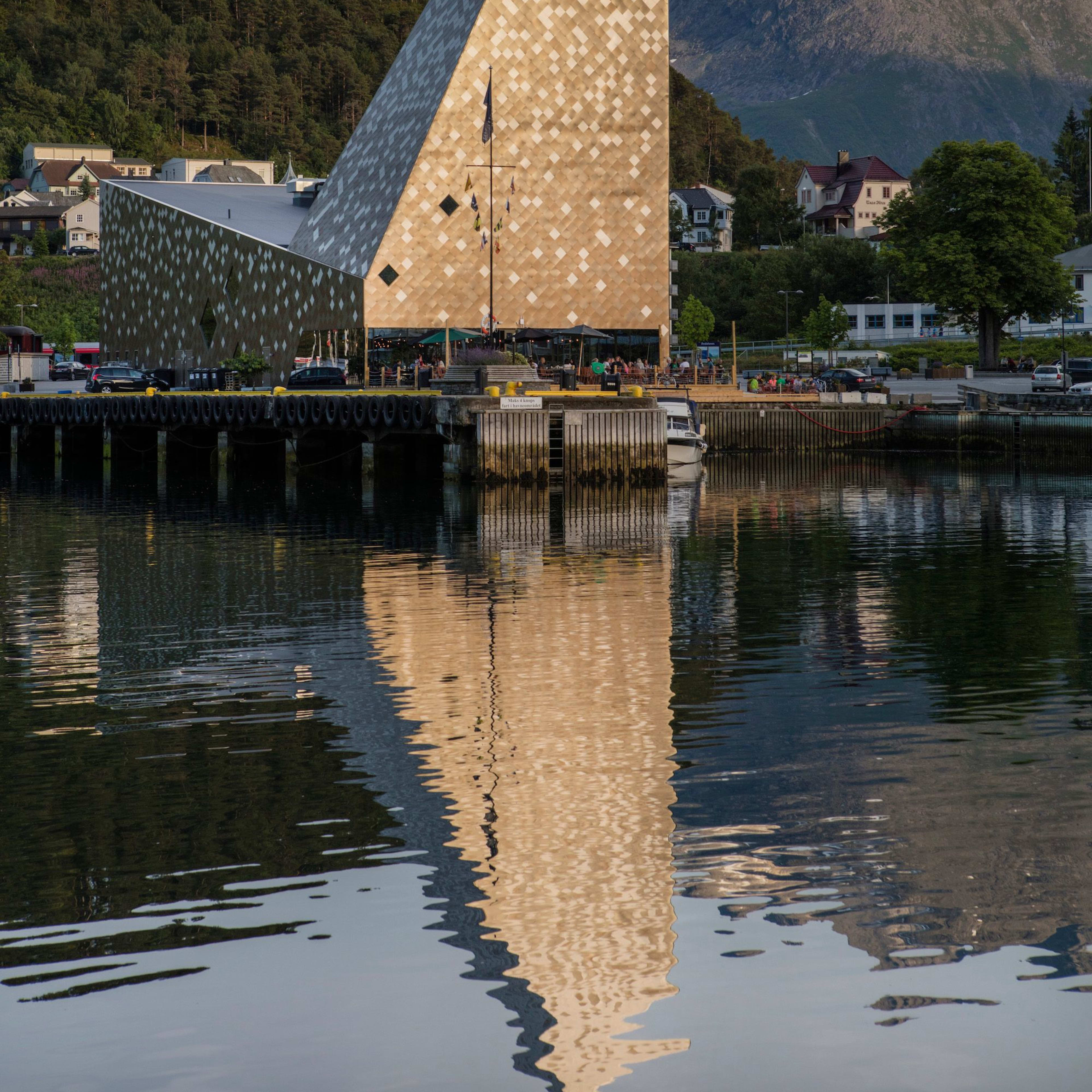 A striking modern building with a pyramid-like shape, located by the water with reflections in the calm water below.