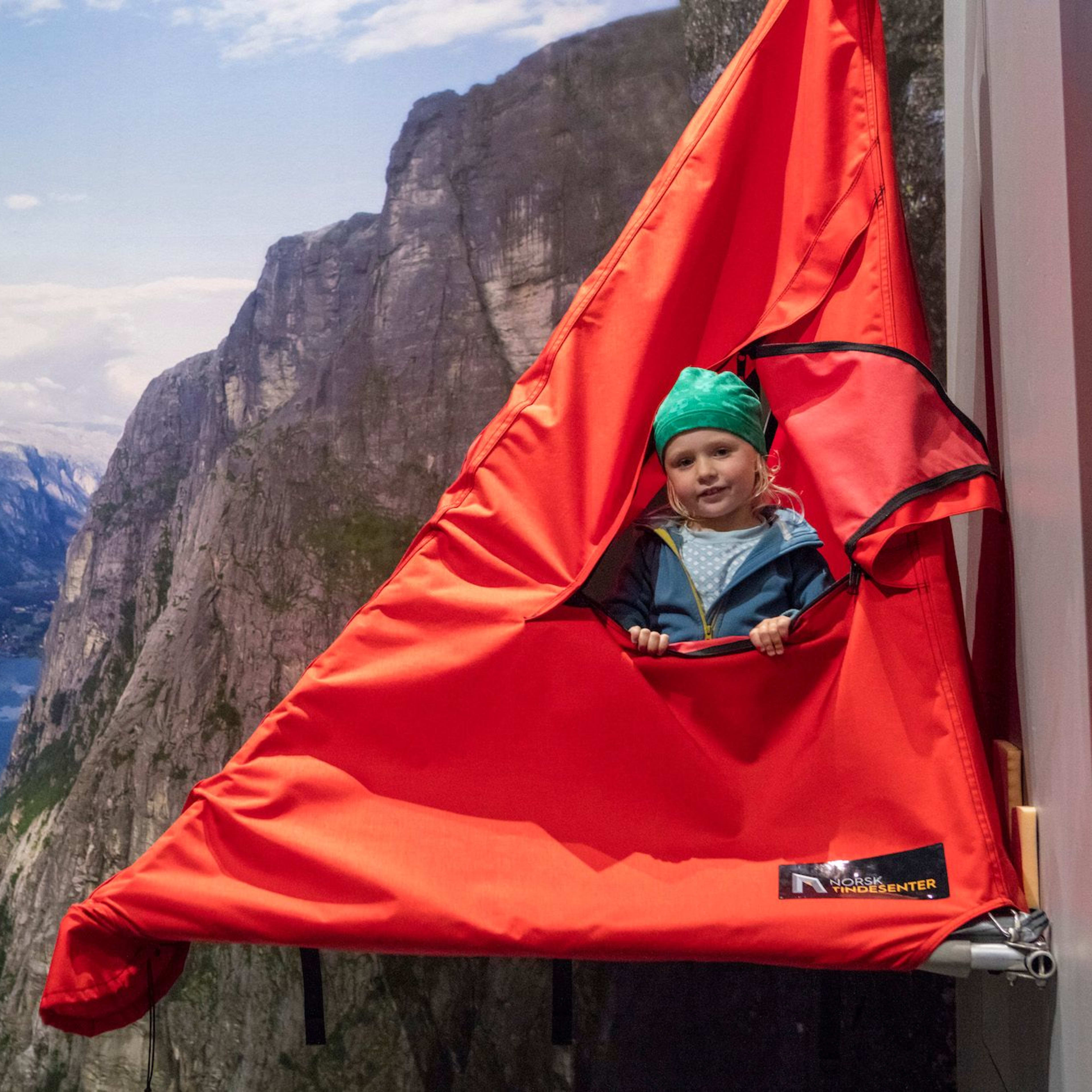 A child sitting in a bright red hanging tent with a photograph of a fjord in the background.