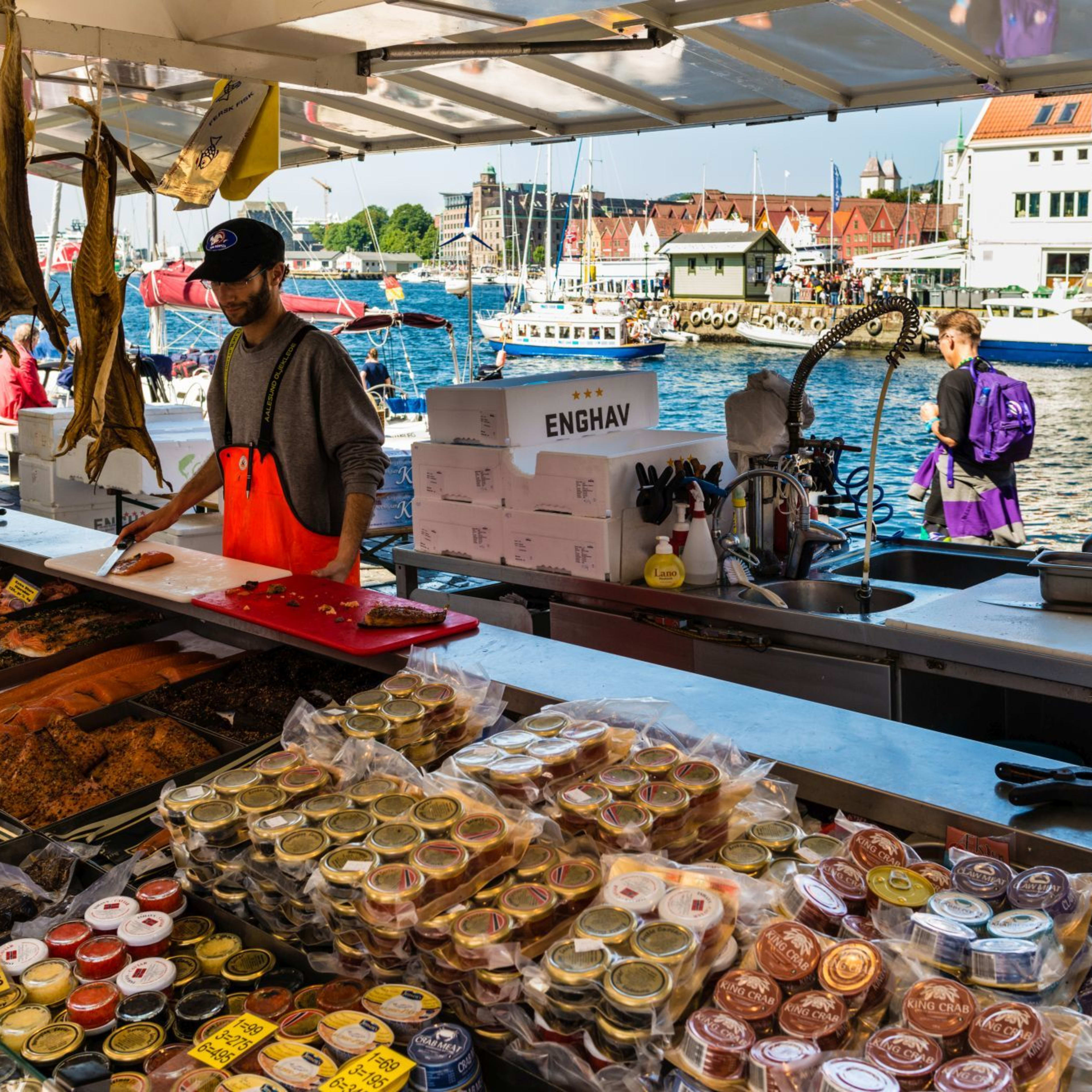 Fishmonger at Bergen market prepares seafood at a stall surrounded by fresh fish, boats, and waterfront buildings.
