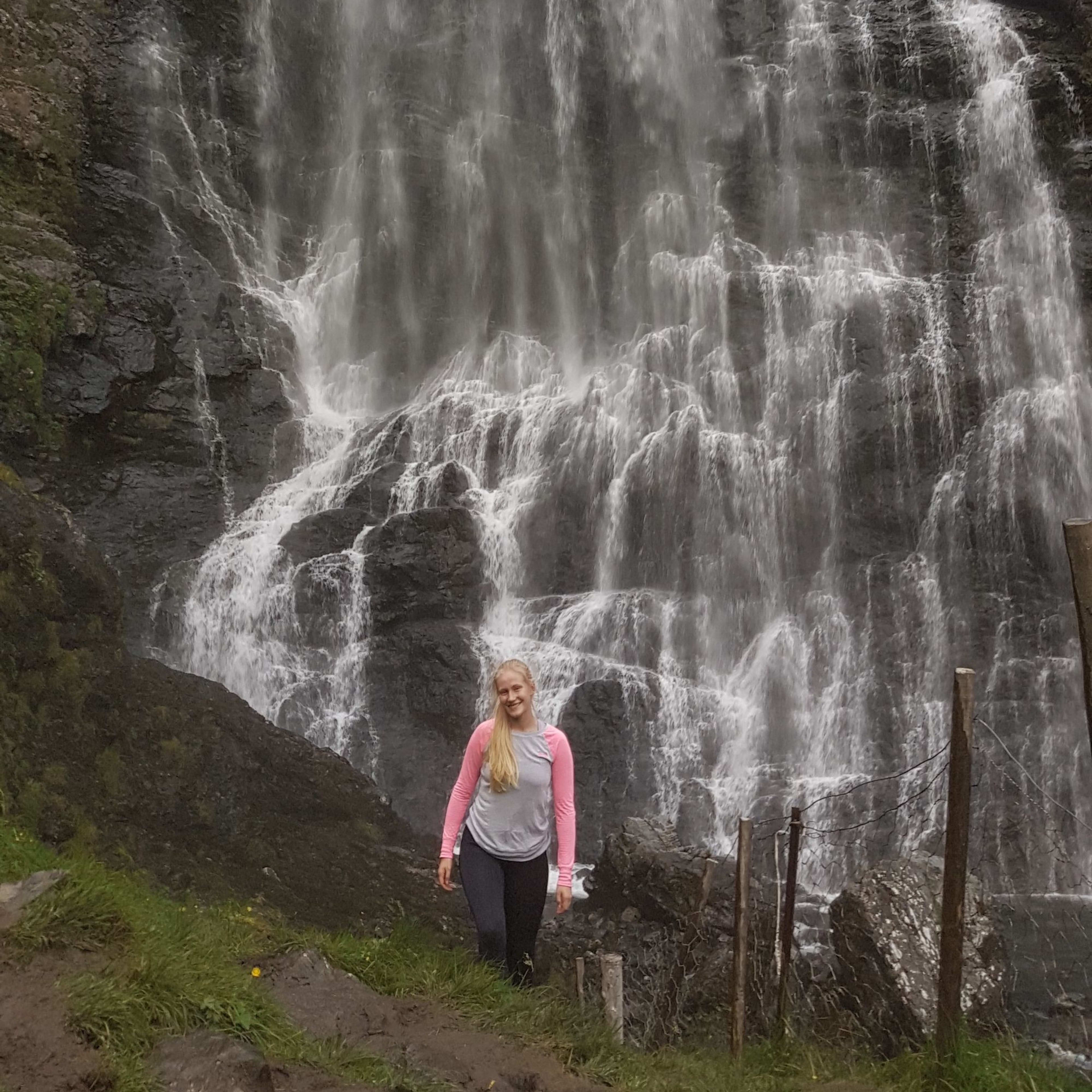 Kvinne smiler ved foten av høy foss.
