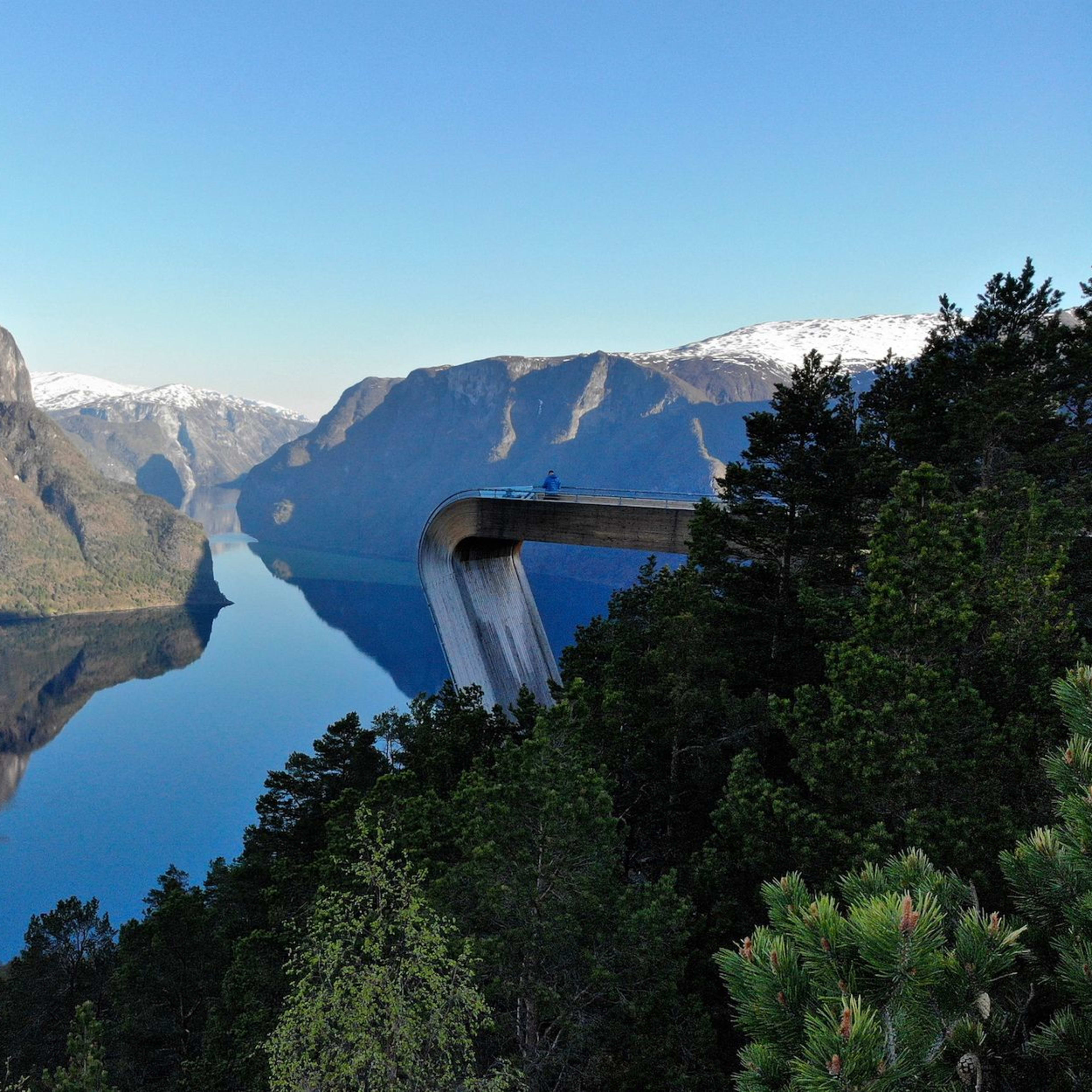 Modern viewpoint juts over fjord, reflected in calm water below, with snowy peaks all around.