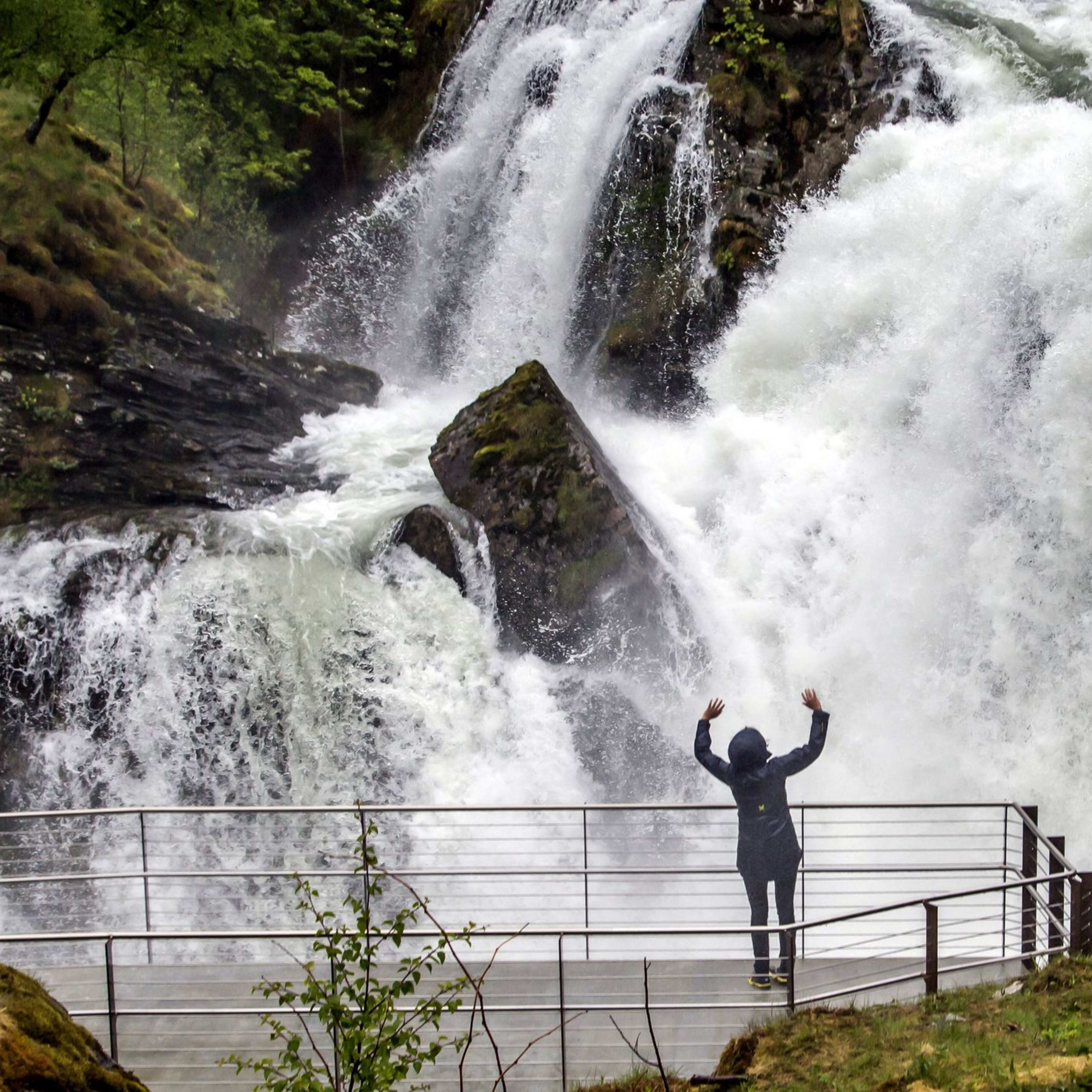 Person in Regenjacke hebt die Arme vor gewaltigem Wasserfall im Grünen.