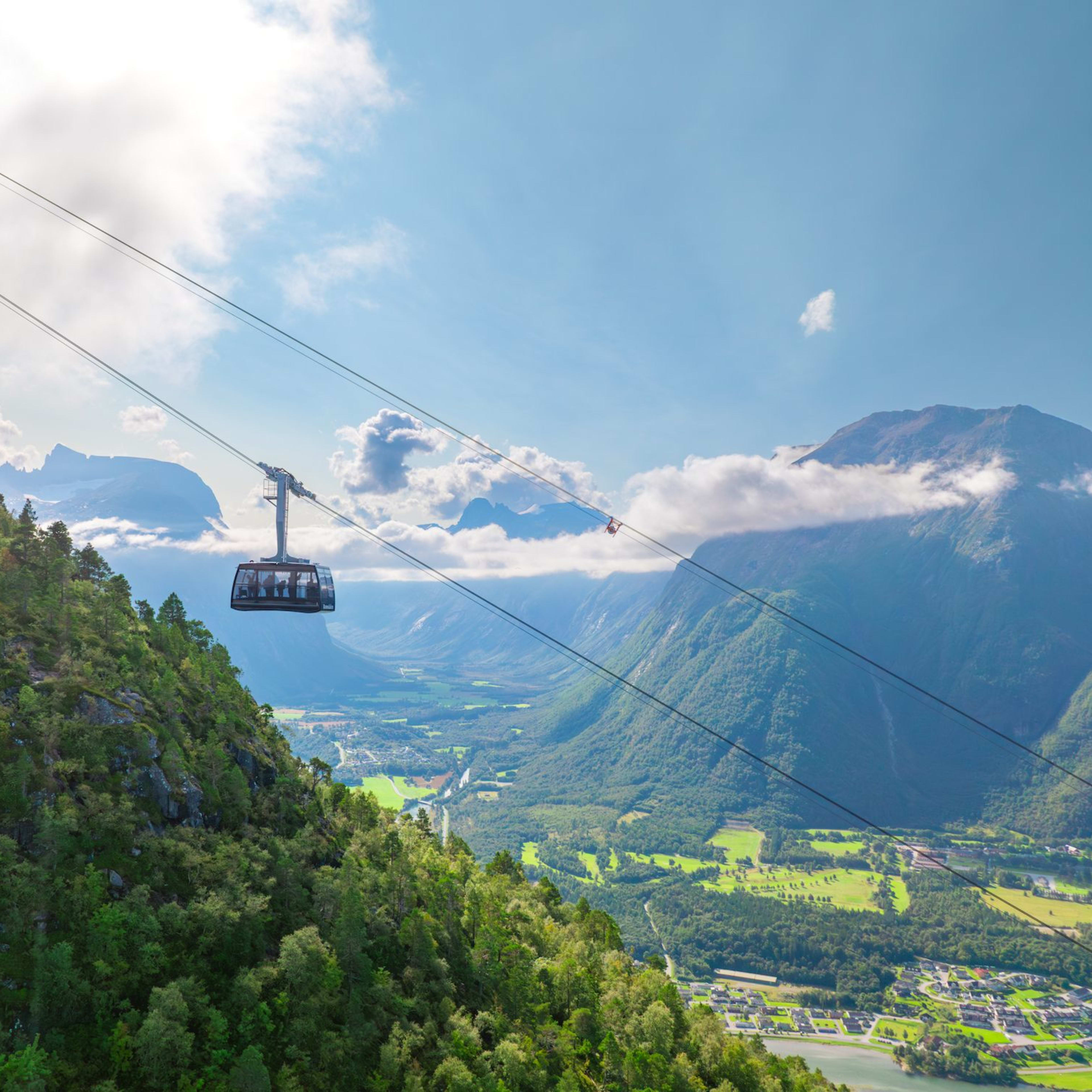 Eine Seilbahn fährt über einen üppig grünen Berg, mit Feldern, Dörfern und gewundenen Straßen darunter und hohen Bergen in der Ferne.