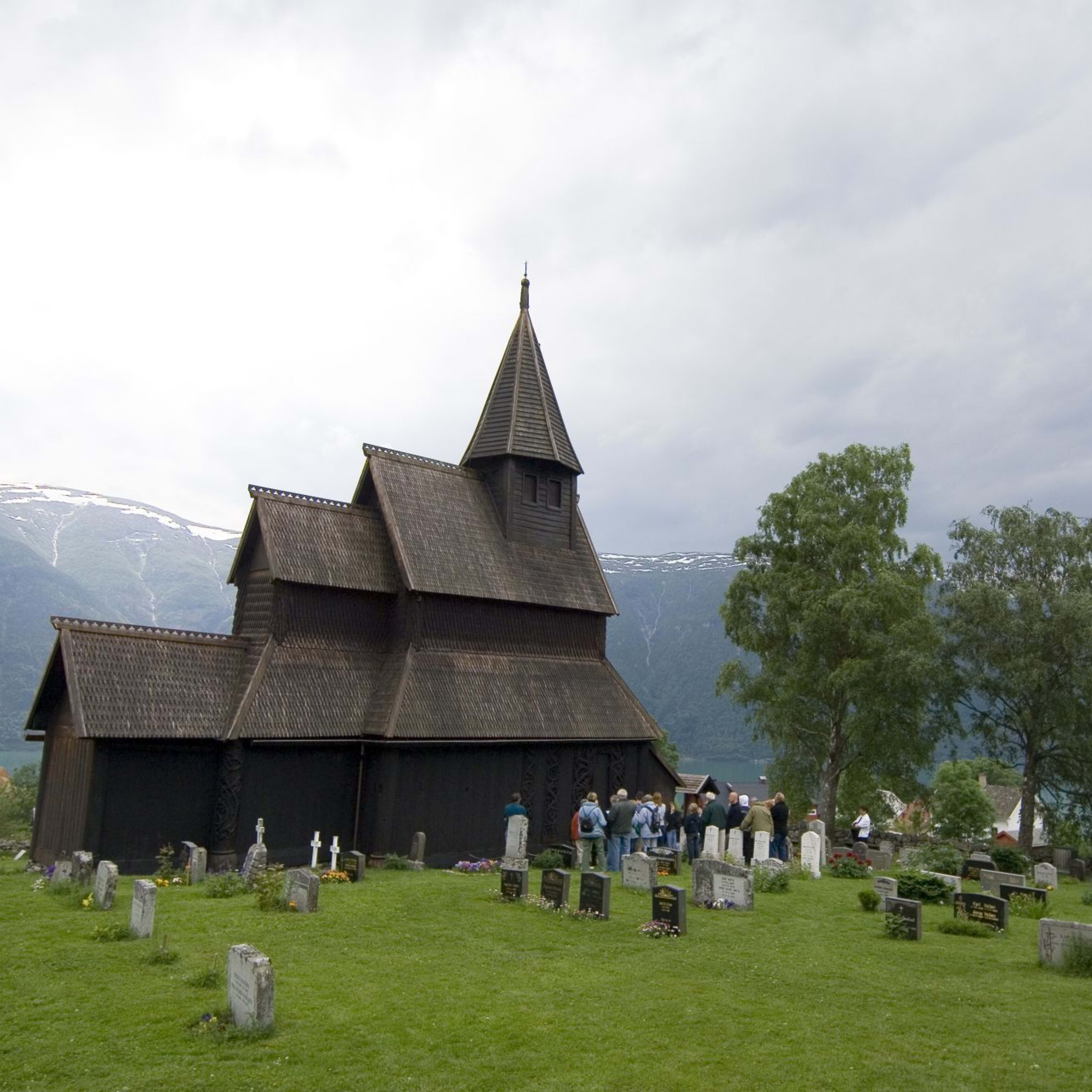Gruppe utenfor stavkirke med grønn kirkegård og fjord i bakgrunnen.