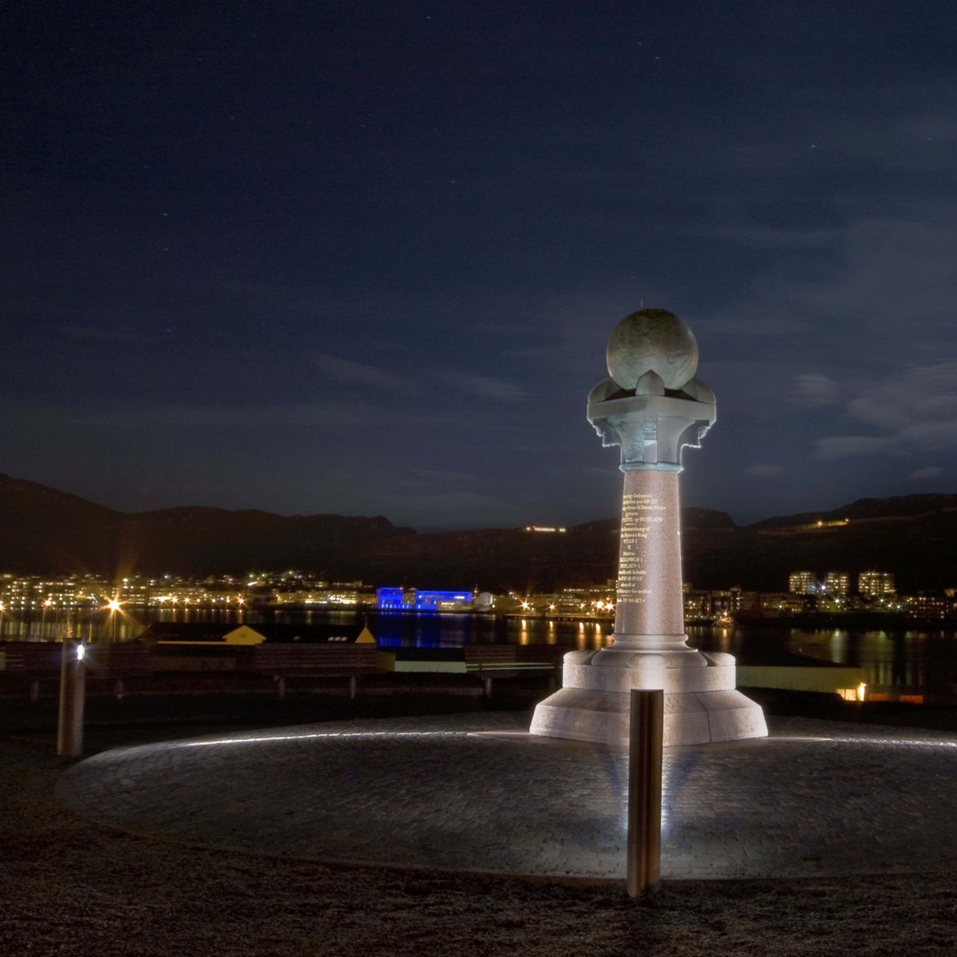 Høyt monument med globus på toppen lyser om natten, med bylys reflektert i vann.