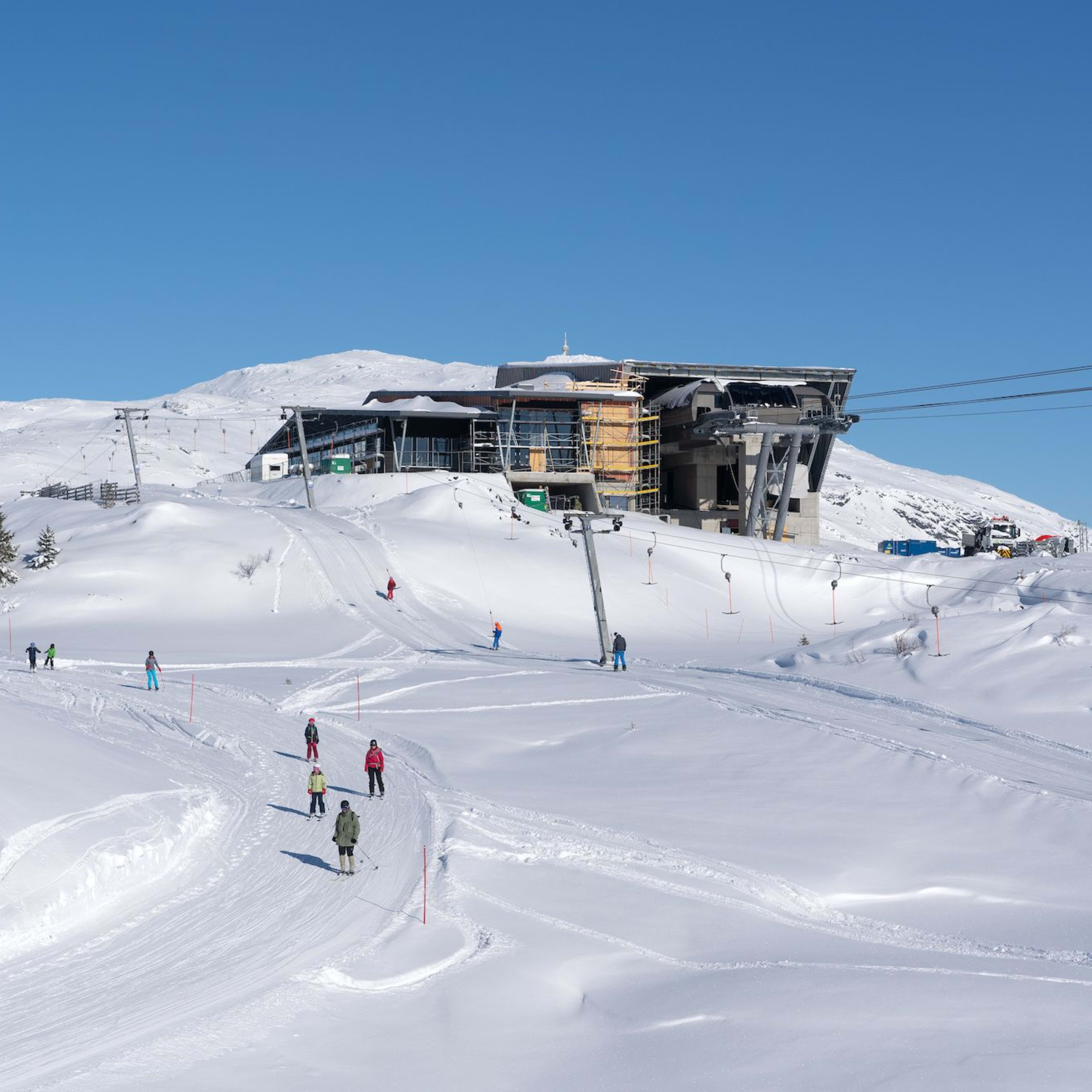 Skiers cross snowy slope near modern cable car station under clear blue winter skies.
