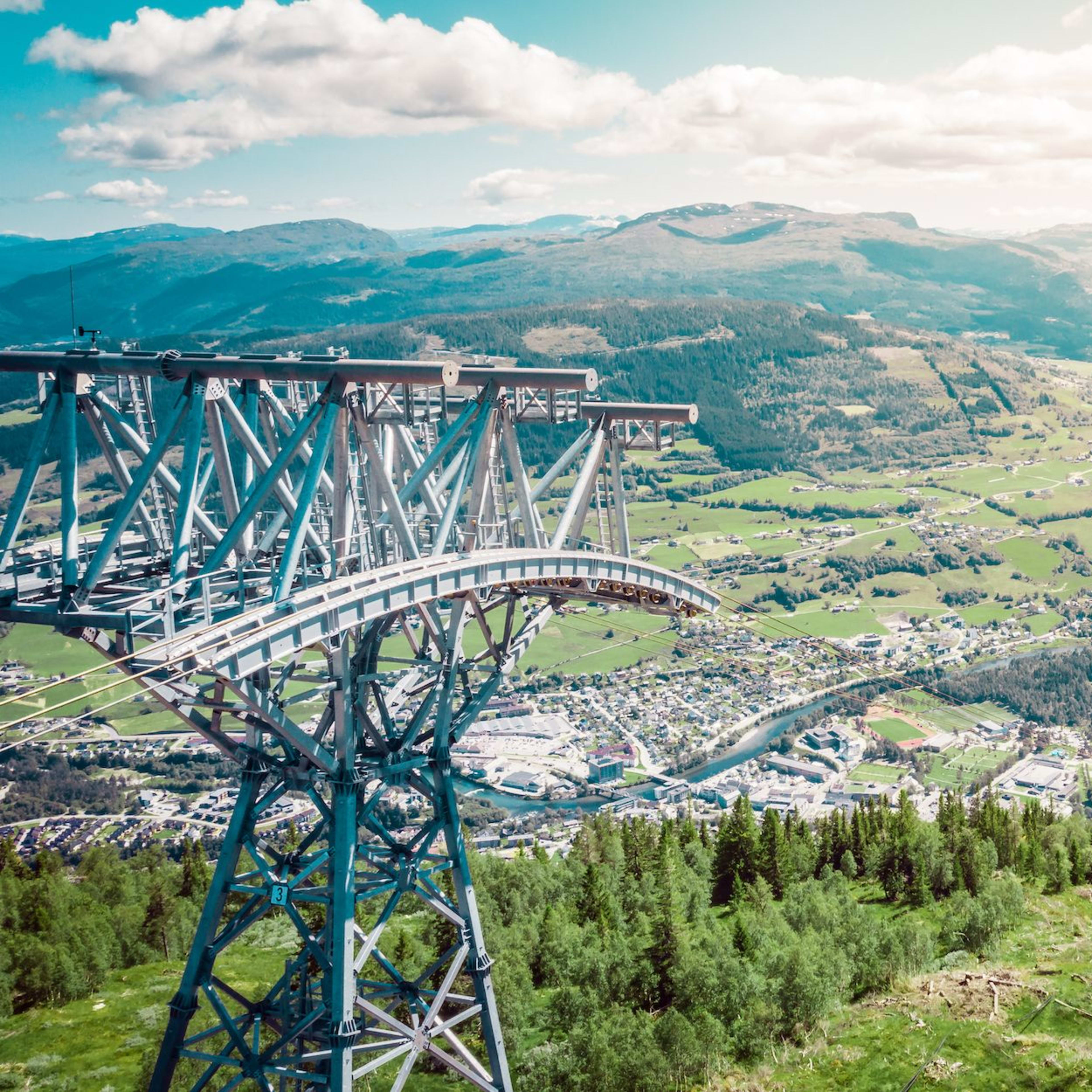Steel tower on green slope overlooks lake, roads, and valley towns under partly cloudy skies.