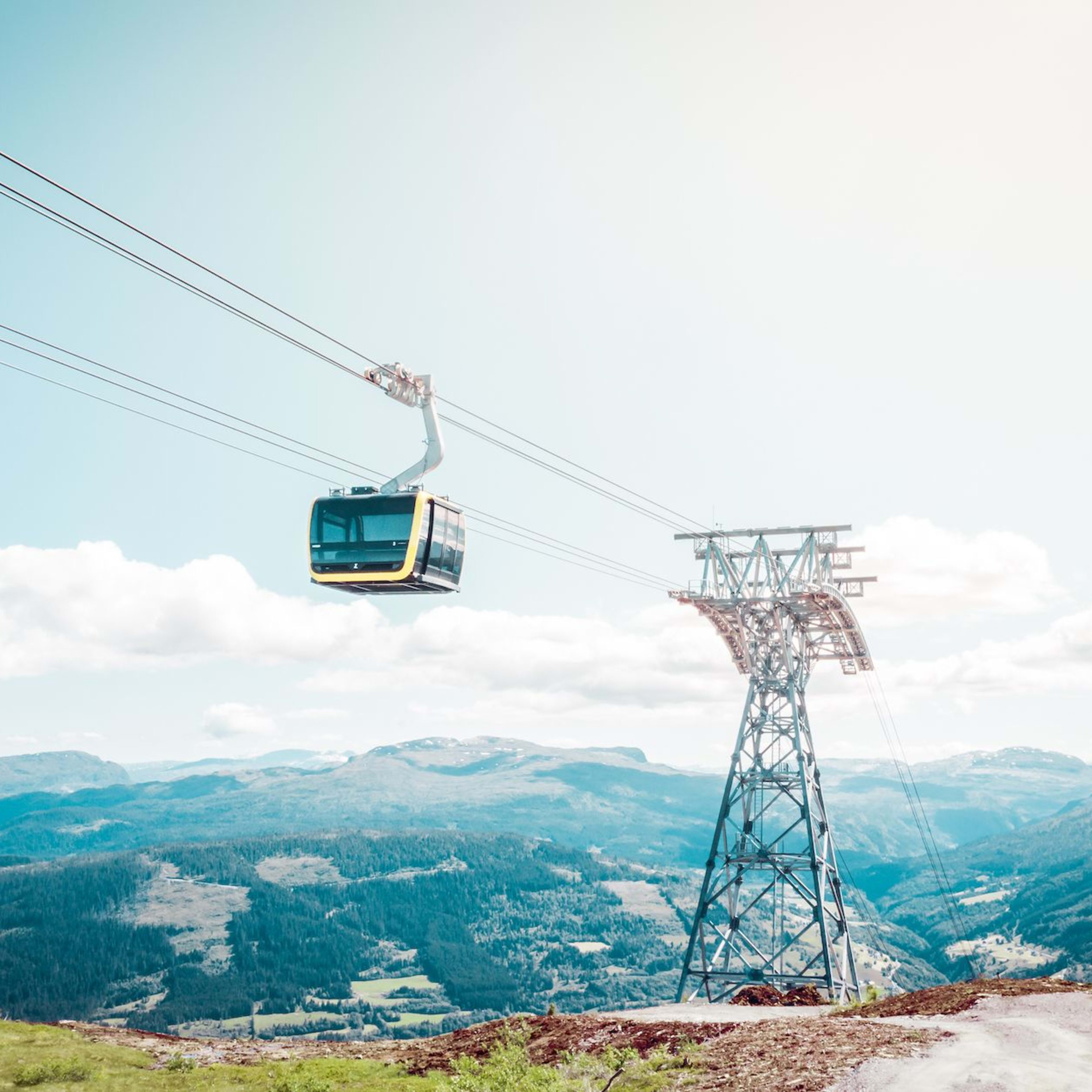 Yellow-black cable car travels high above green valleys and peaks under bright clear sky.