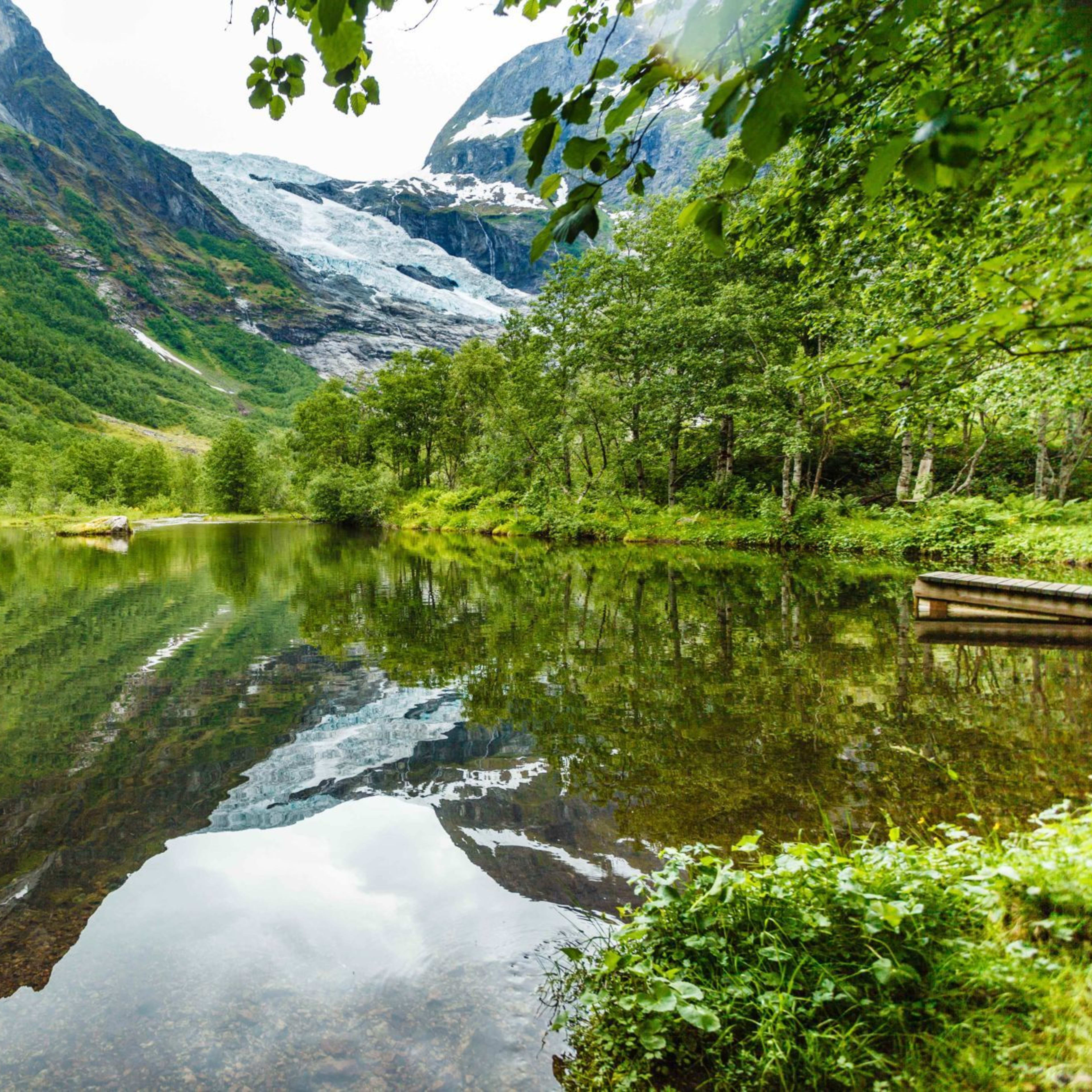 Fjellvann speiler isbre og skog, brygge til høyre.