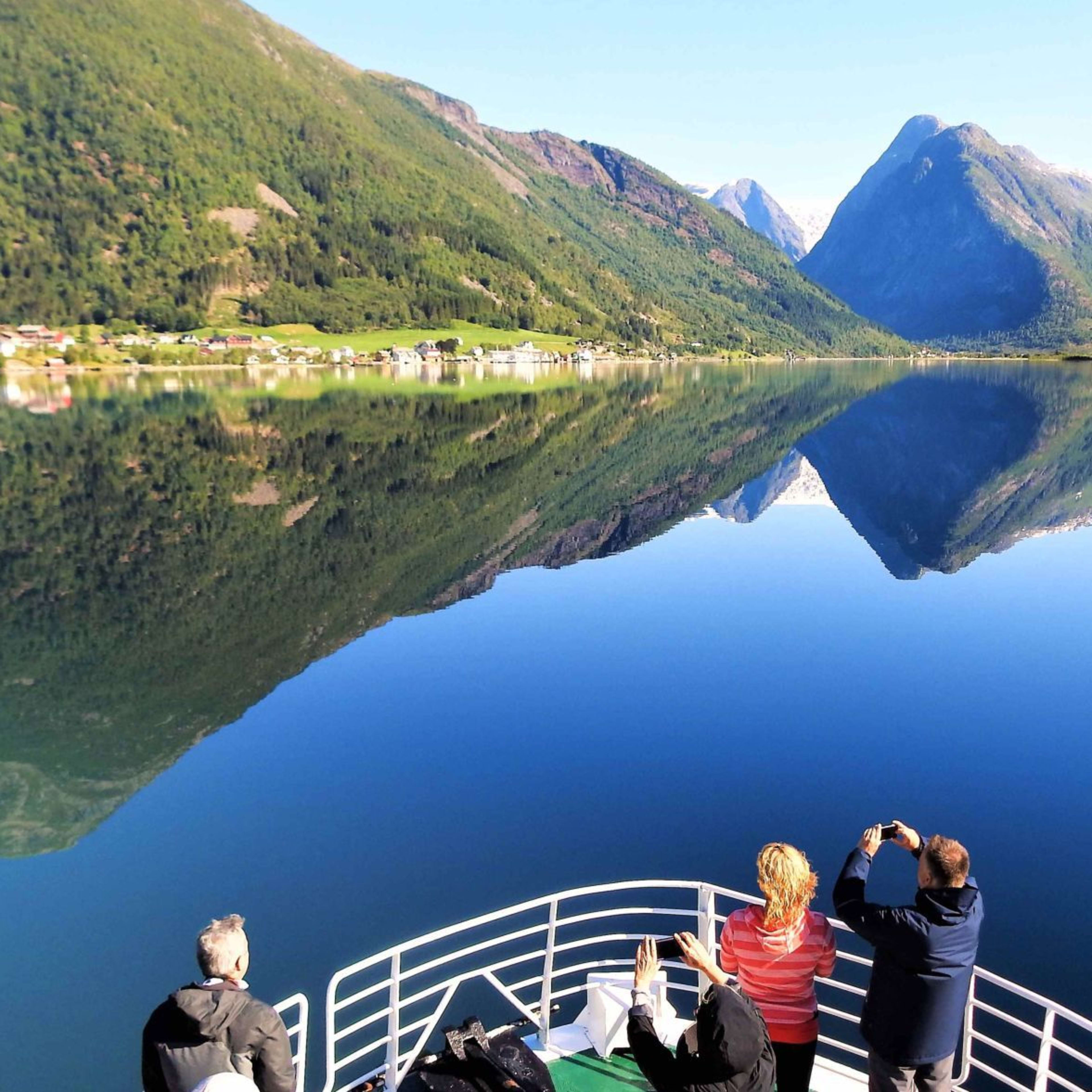 Turister nyter speilblank fjord med snødekte fjell i det fjerne.