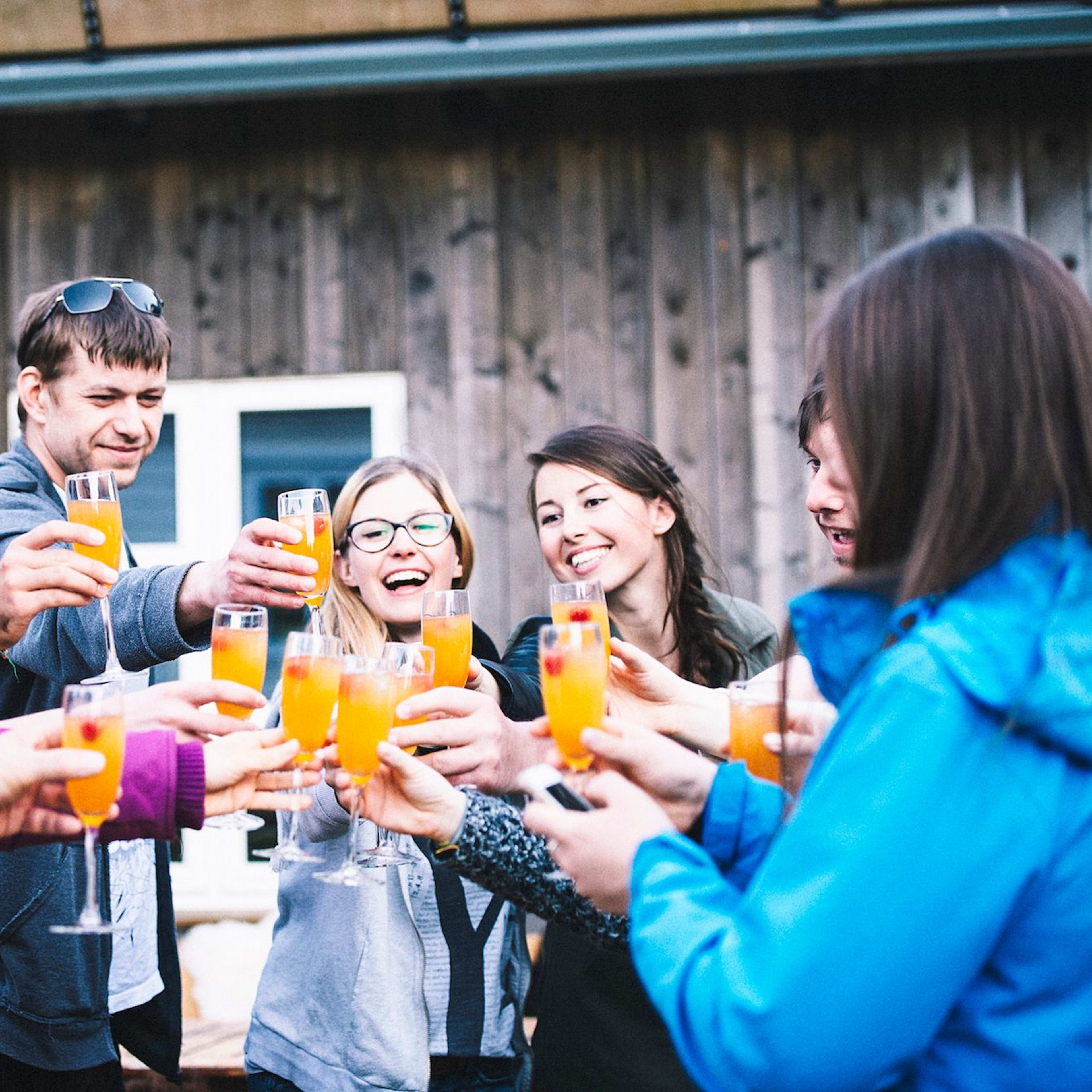 Freunde stoßen draußen mit Orangendrinks vor Holzhaus fröhlich an.