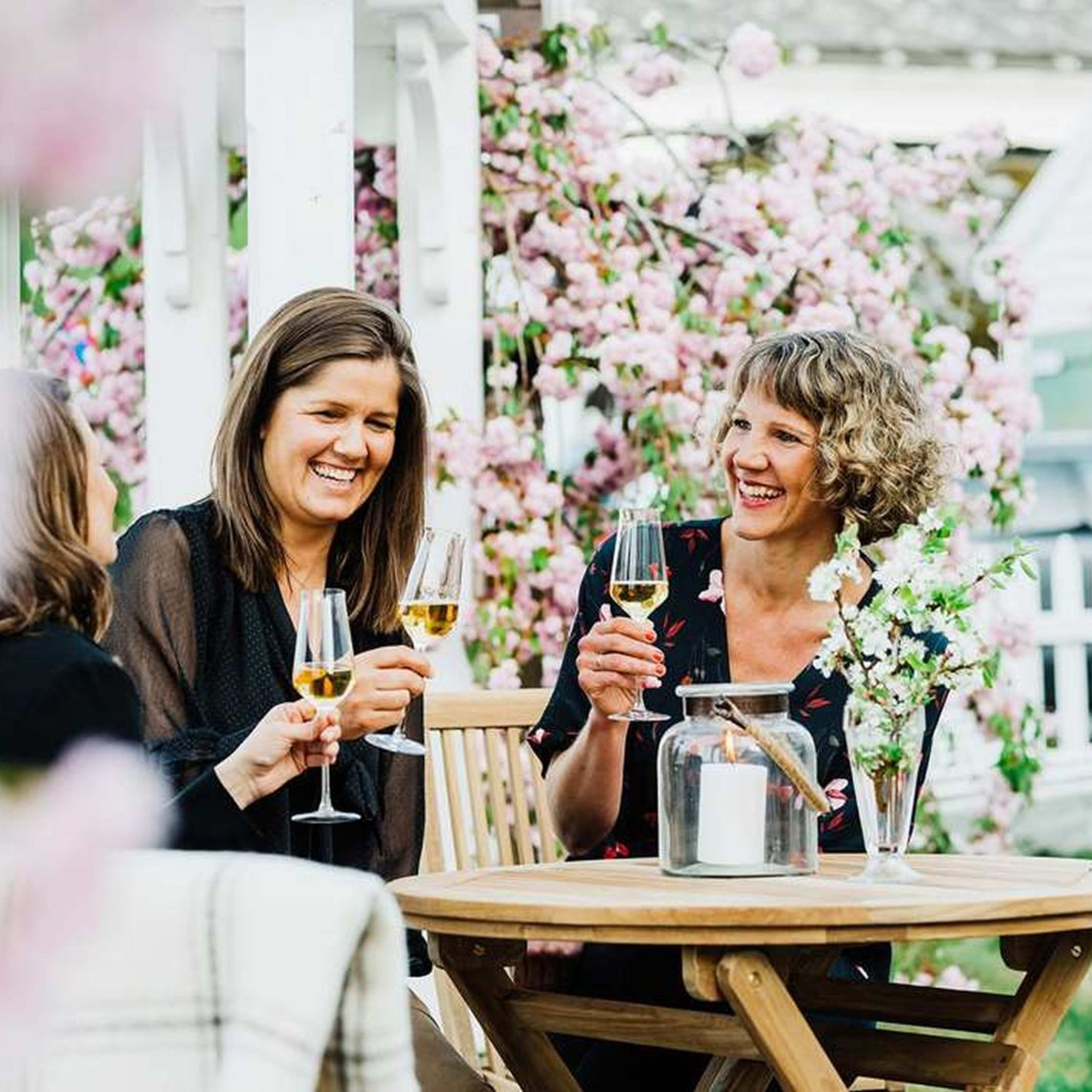 Women laugh and enjoy drinks cider in flower-filled garden in Ulvik.