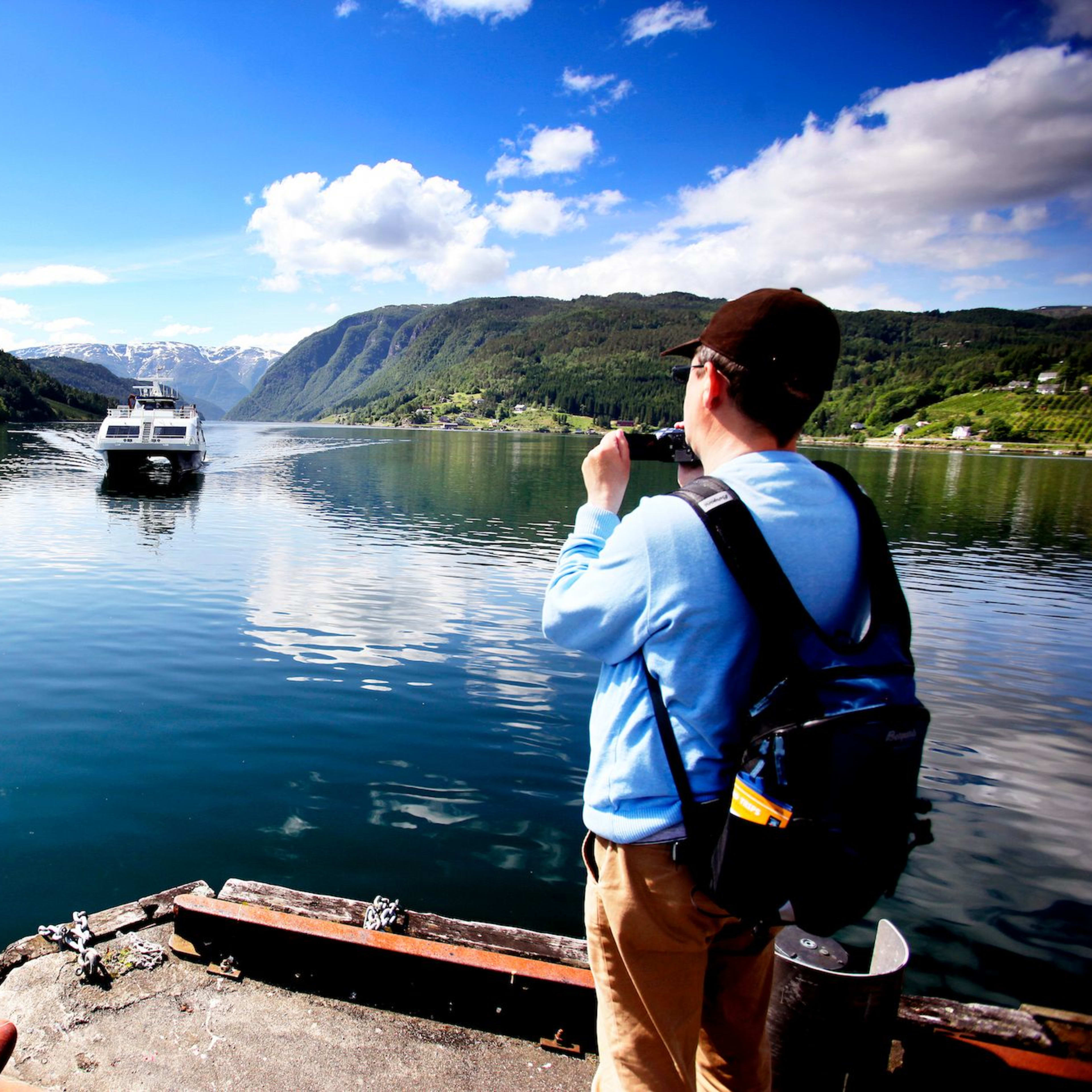 Person fotografiert Boot auf ruhigem Hardangerfjord von Steg mit Bergen im Hintergrund.